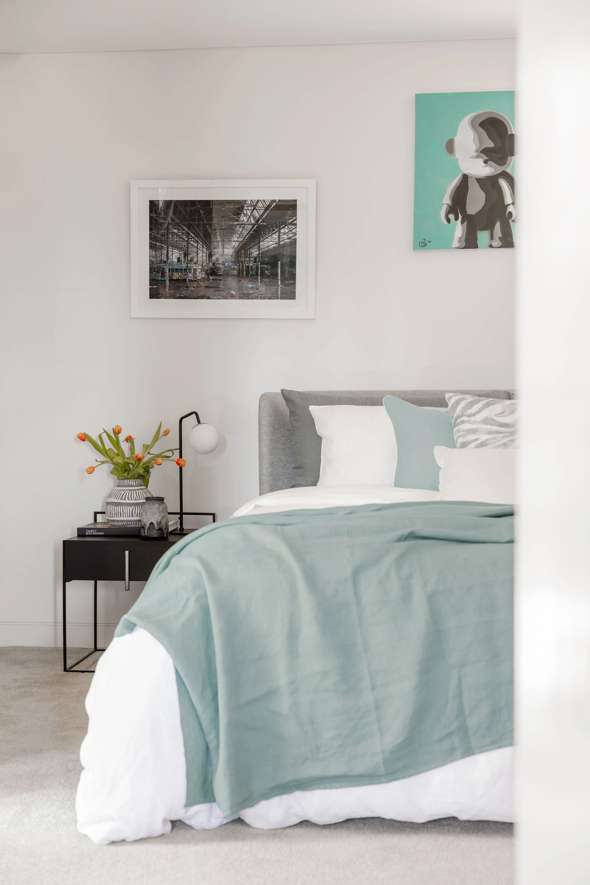 A bedroom with a bed covered in white and light blue bedding, a black nightstand with a striped vase of orange tulips and a round white lamp, artwork on the wall including a black-and-white photograph of an industrial interior and a canvas with a sty