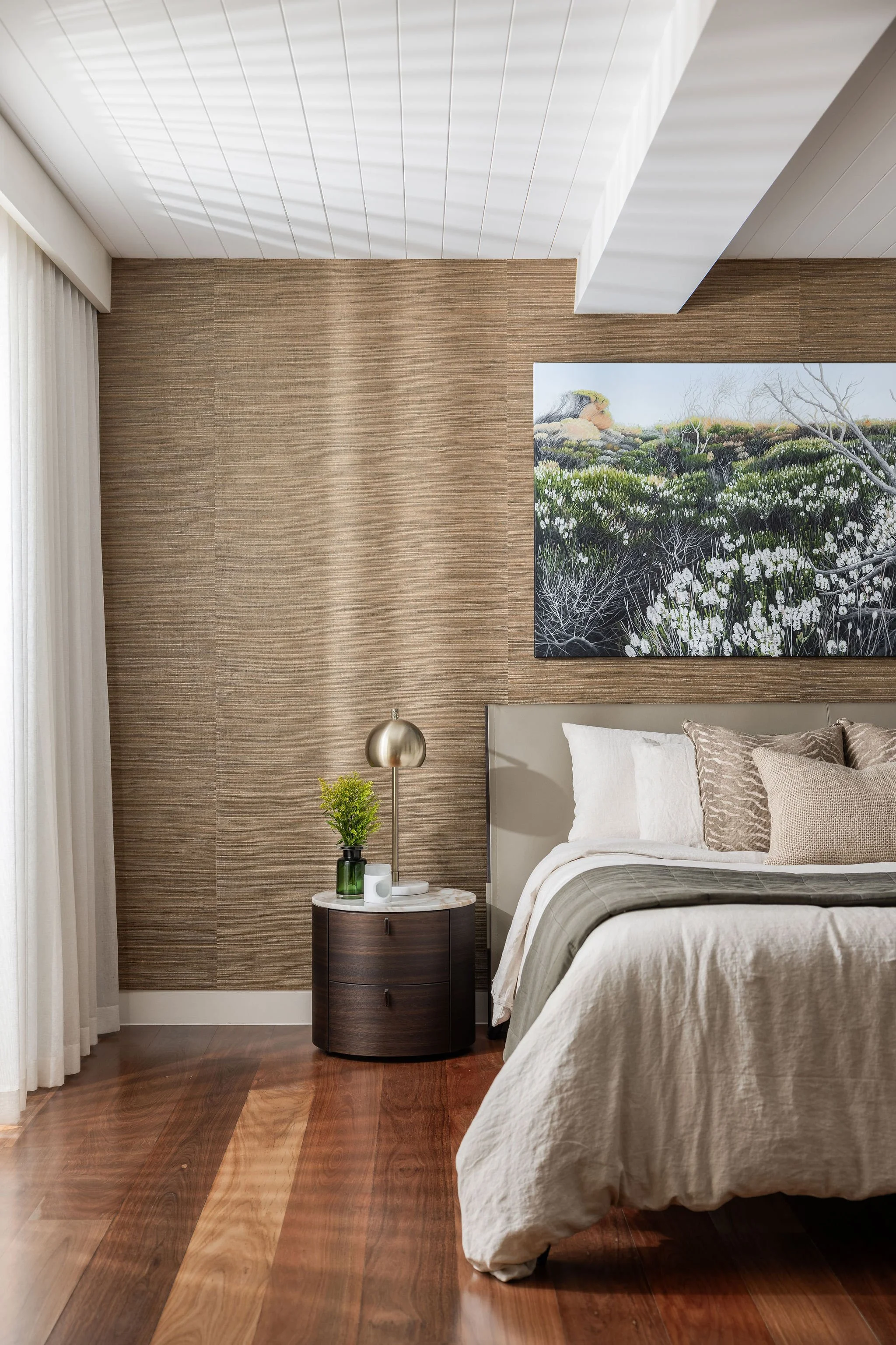 A bedroom with a bed covered in beige and white linens, a round dark wood nightstand with a small potted plant, a brass table lamp, and candles. The wall features a large landscape painting, and the floor is made of polished wood. Light-colored curta