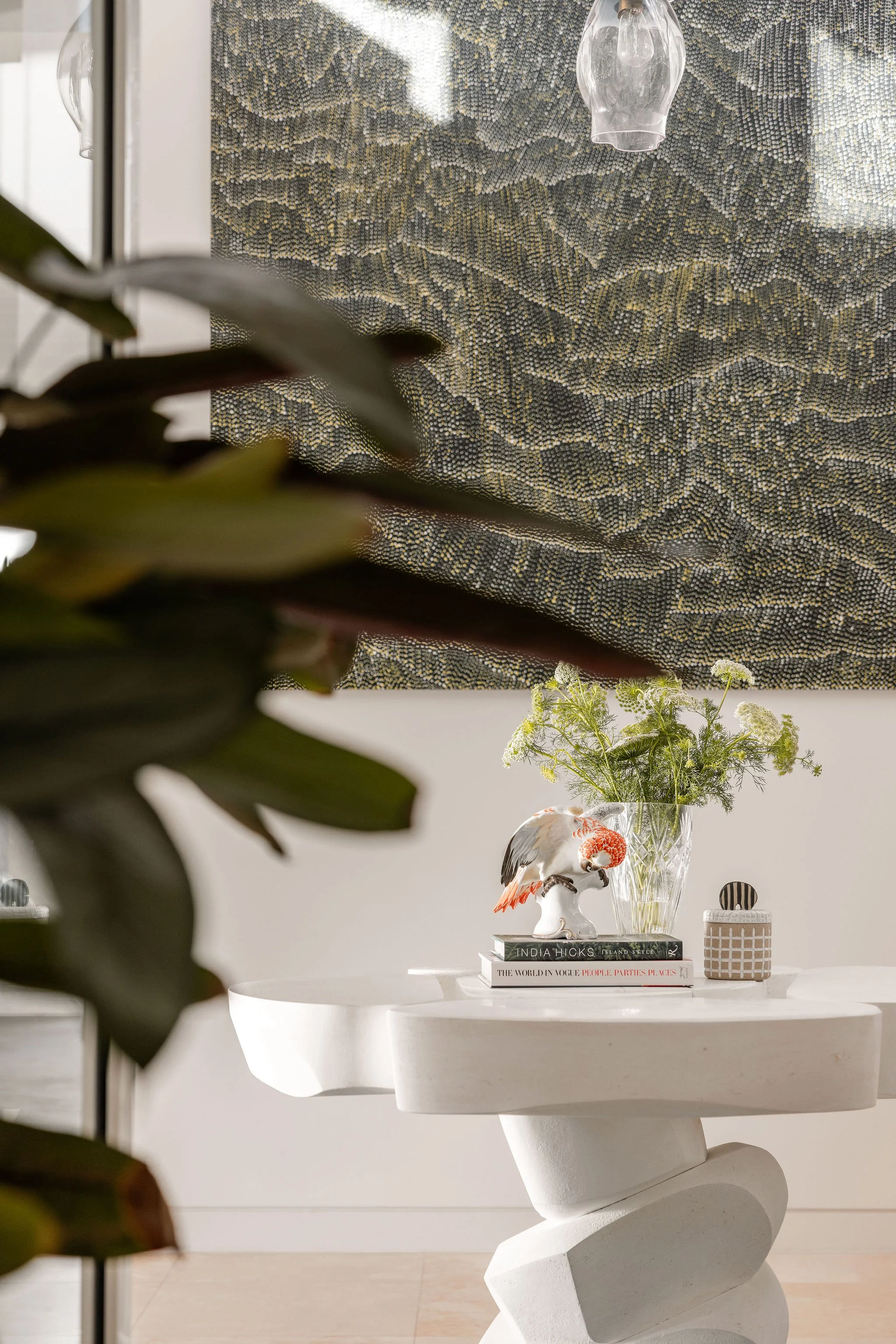 A white round table with books, decorative objects, and a glass vase with green plants and white flowers. A ceramic bird figurine is placed on top of the books, which include titles such as 'India Hicks' and 'The World in Vogue.' Part of a dark plant