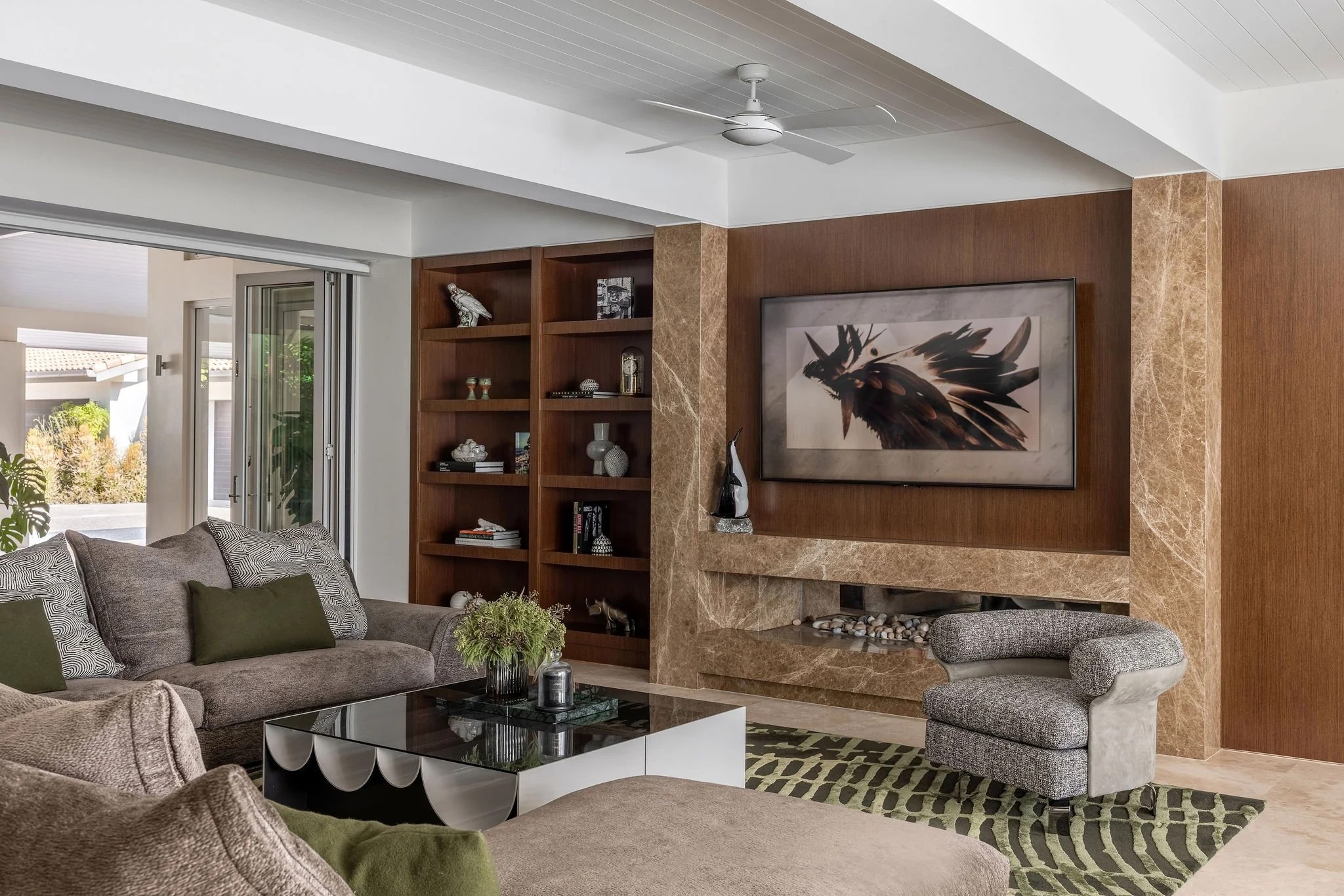 Gold Coast Living room with Jardan Vista sectional sofa, green pillows, glass coffee table, and a built-in wooden shelf. Large framed artwork of a bird is on a brick wall with a fireplace below. There are sliding glass doors leading outside.