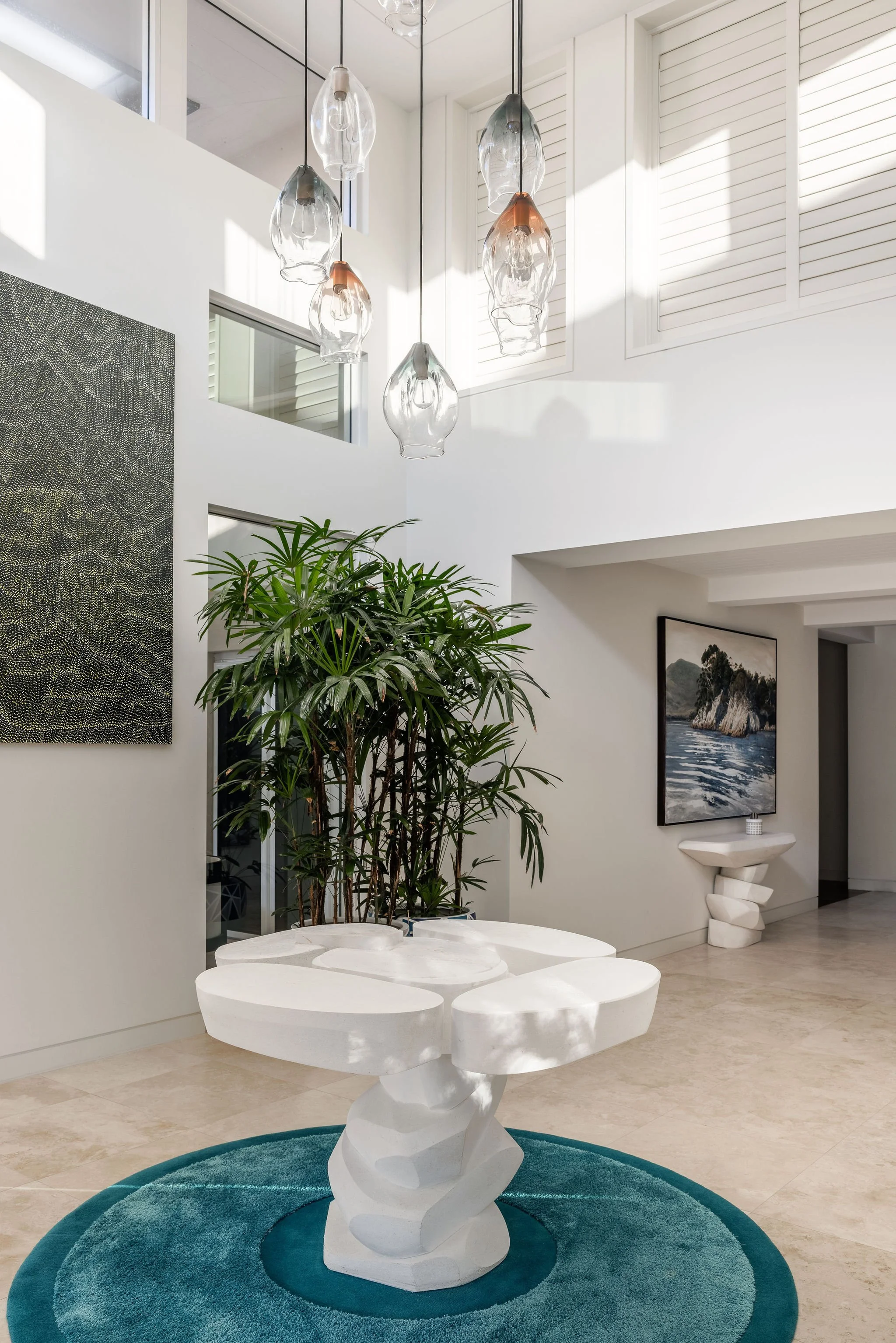 Modern white sculpture resembling a twisted, abstract form placed on a teal circular rug in a bright, spacious room with white walls, a large green plant, artwork, and contemporary lighting fixtures.