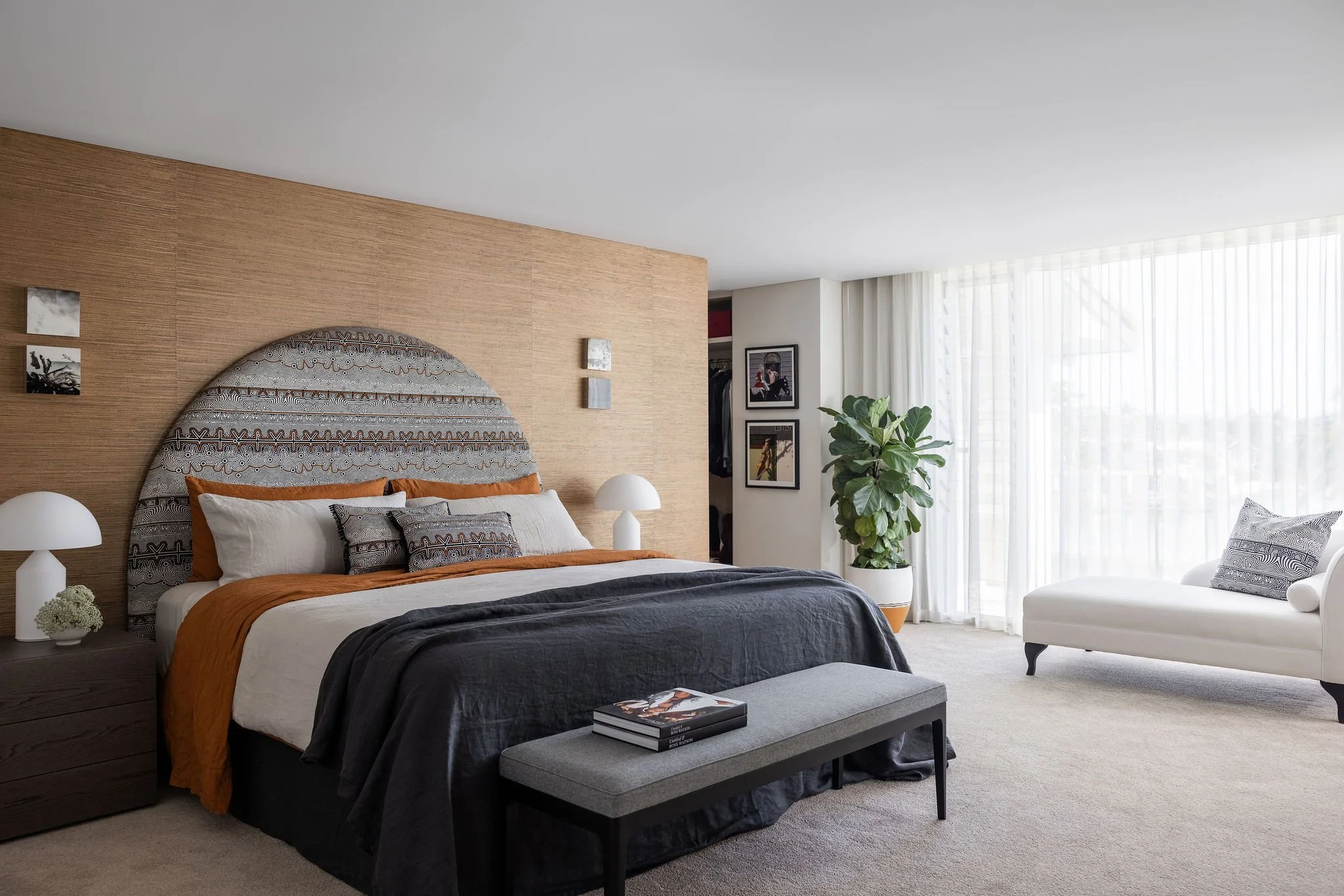 A modern bedroom with a large bed featuring a grey, black, and orange bedding set, a wooden headboard, white side tables with white lamps, artwork on a wooden accent wall, a white armchair with patterned pillows, a large potted plant, and sheer white