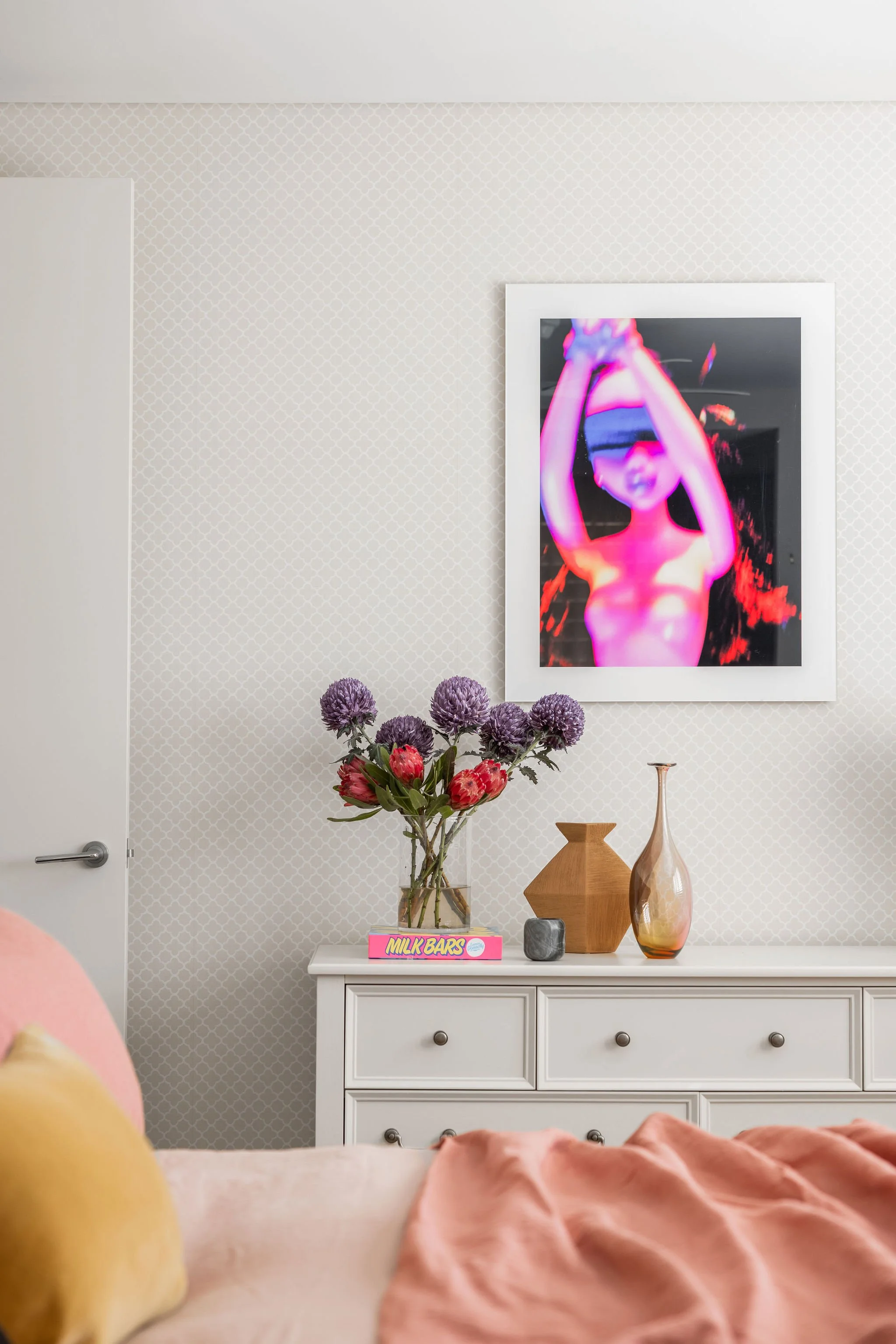 A bedroom scene seen from a bed with a yellow pillow and a pink blanket. A white dresser with a glass vase of purple and pink flowers, a wooden decorative object, a small gray stone, and a box of Milk Bars is against the wall. Above the dresser hangs
