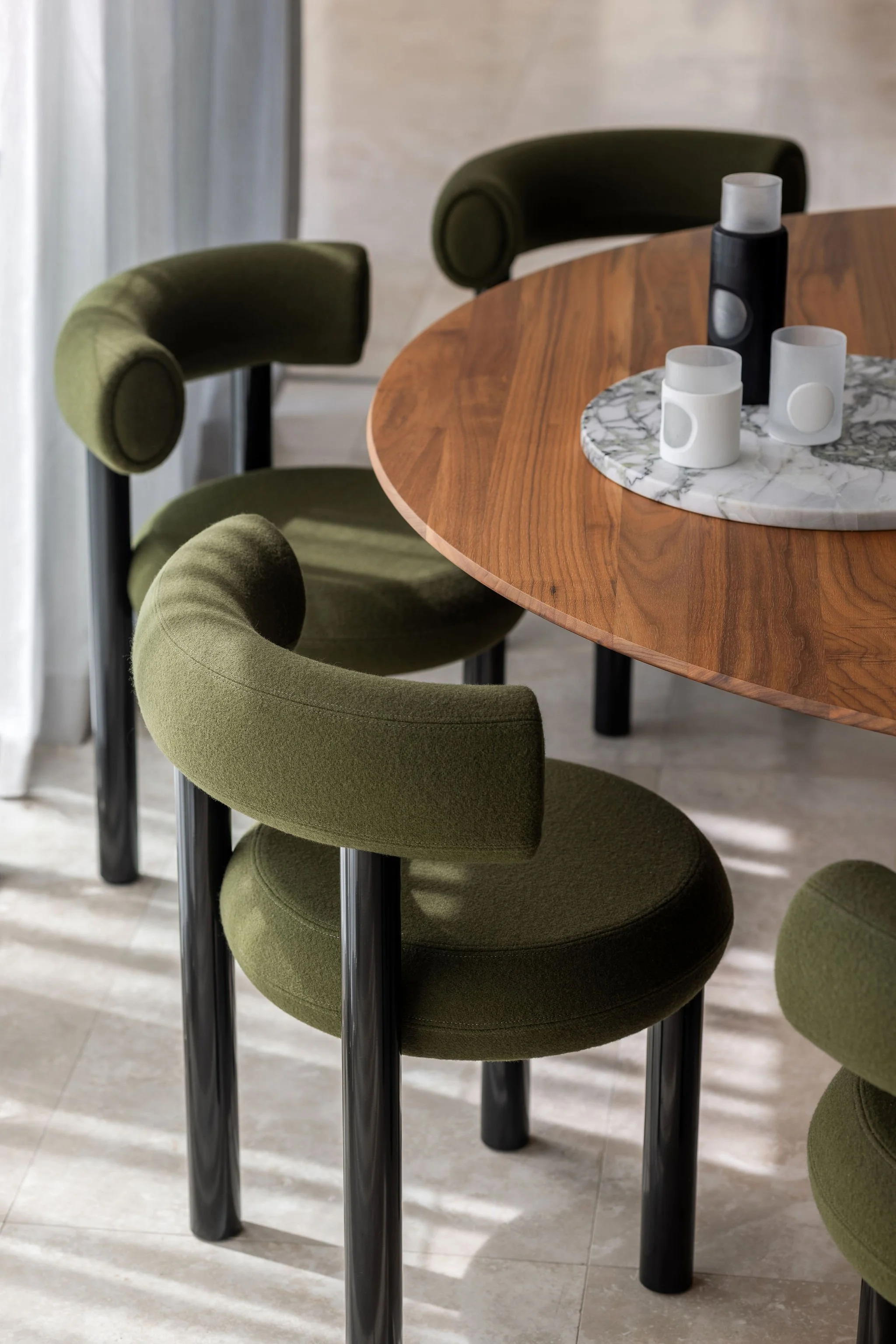 Green upholstered bar stools with black legs lining a wooden round table with a marble centerpiece, in a modern interior setting.