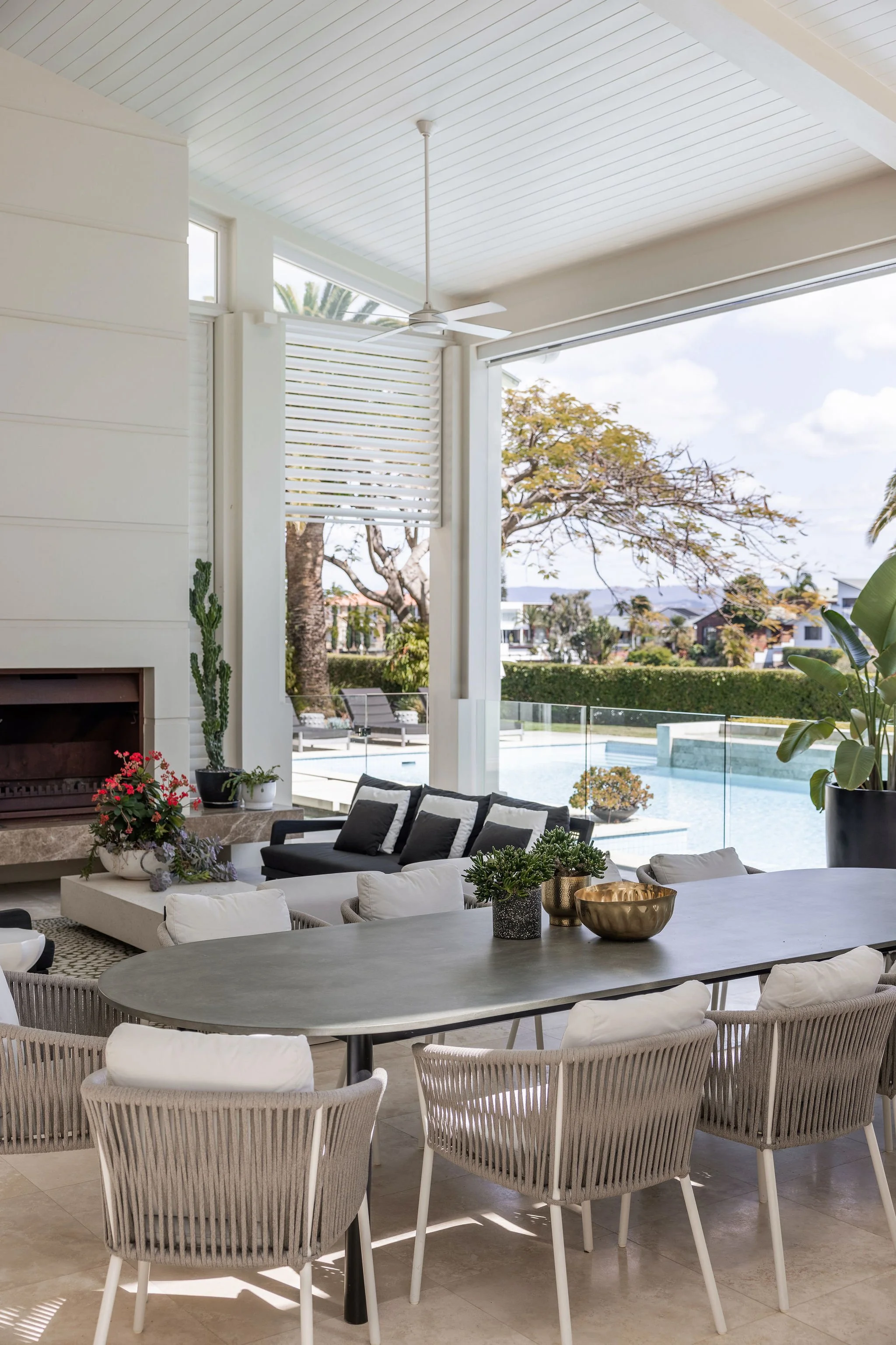 View of a modern indoor living area with large glass windows open to a backyard with a pool, outdoor lounge chairs, trees, and houses in the background. The indoor space features a round dining table with white cushioned chairs, a fireplace, and deco