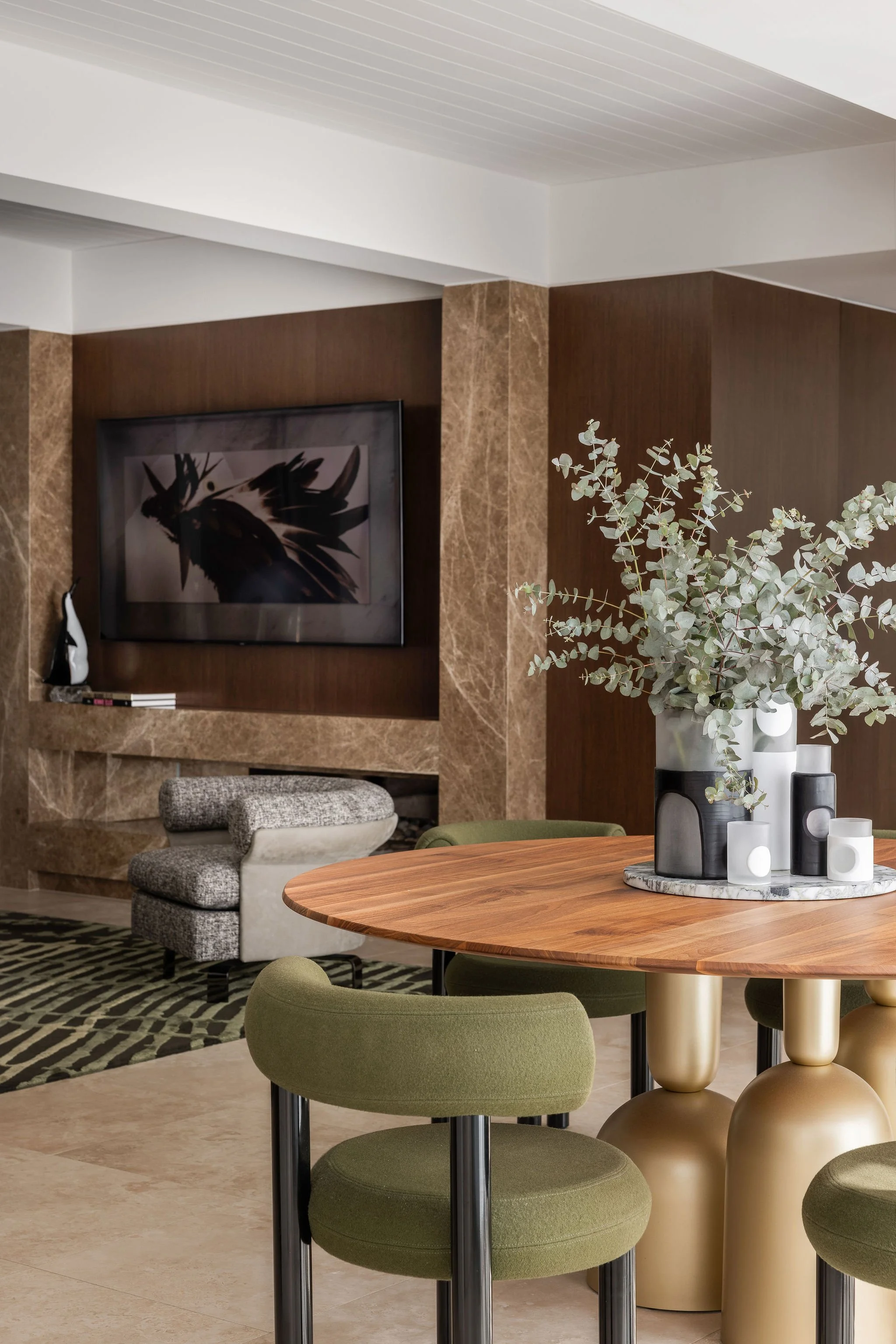 Modern Gold Coast living room with a wall-mounted TV, abstract artwork, plush gray chair, green cushioned stool, round dining table, green upholstered chairs, decorative white and black vases with eucalyptus, and a patterned area rug.