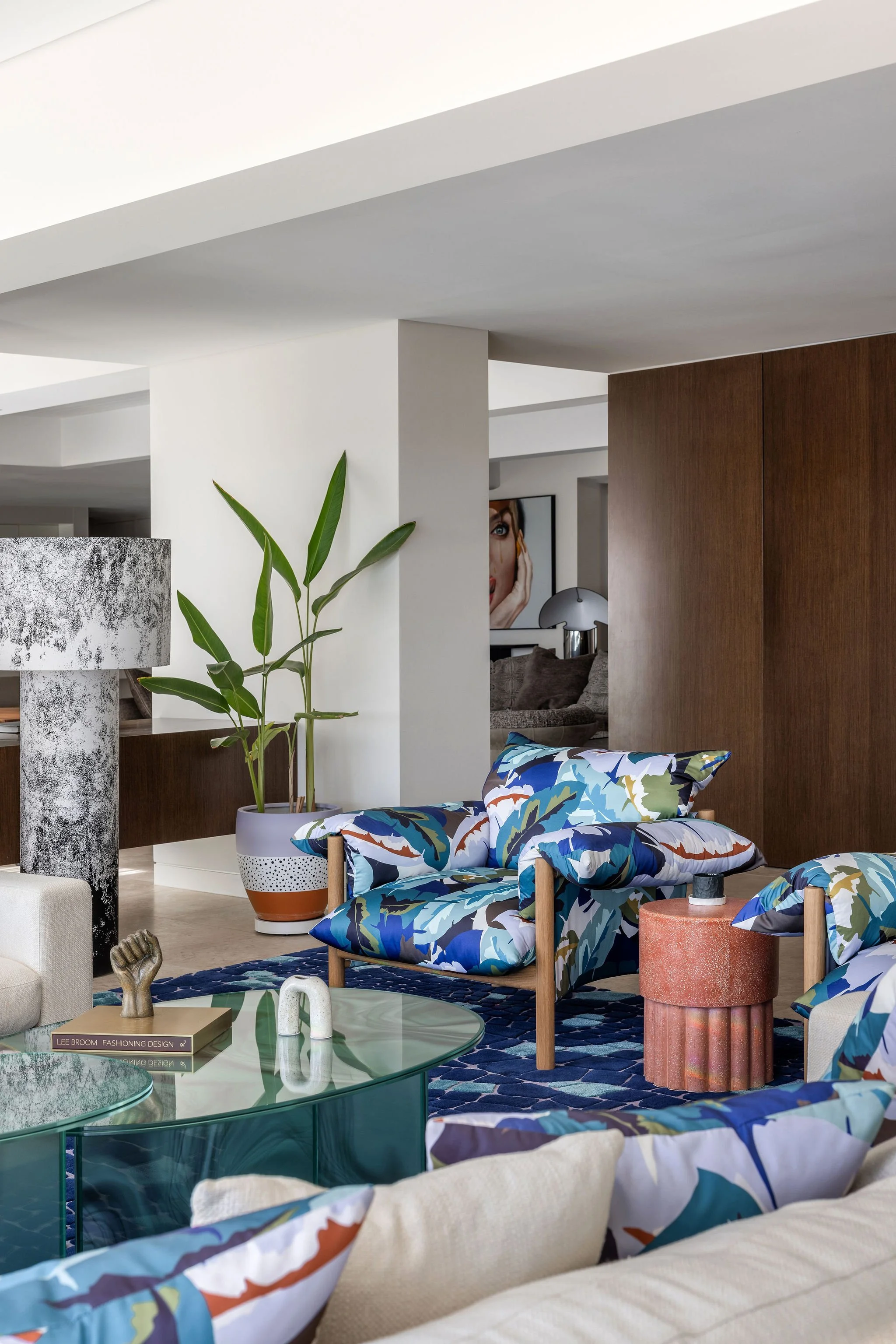 Modern living room with white walls, wood accents, and various colorful furnishings including a sofa with vibrant patterned pillows, a green potted plant, a coral-colored side table, glass coffee tables, and artwork on the walls.