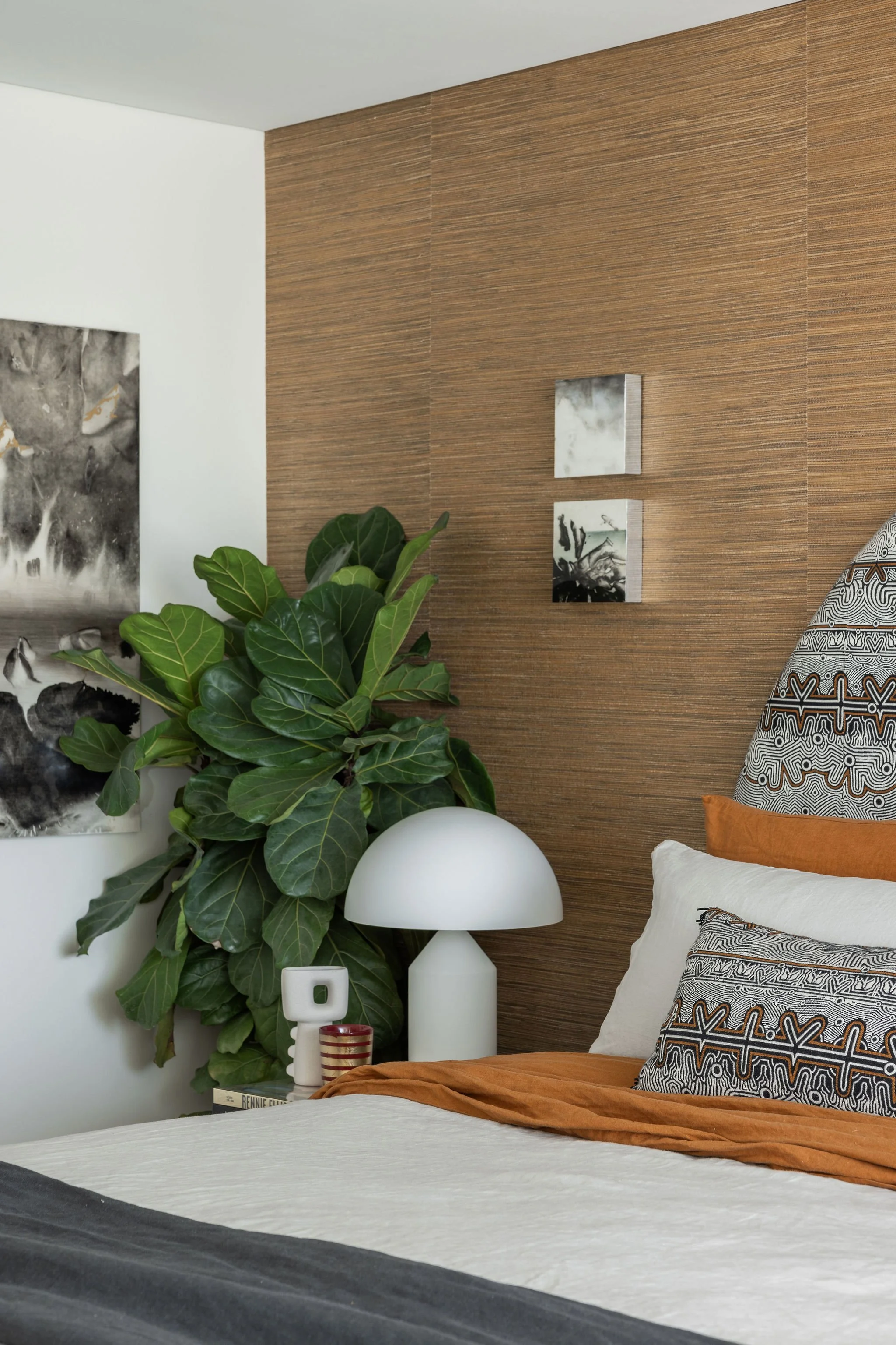 A neatly made bed with an orange blanket and patterned pillows, a white bedside lamp, a potted fig plant, and black-and-white artwork on a wooden accent wall in a modern bedroom.