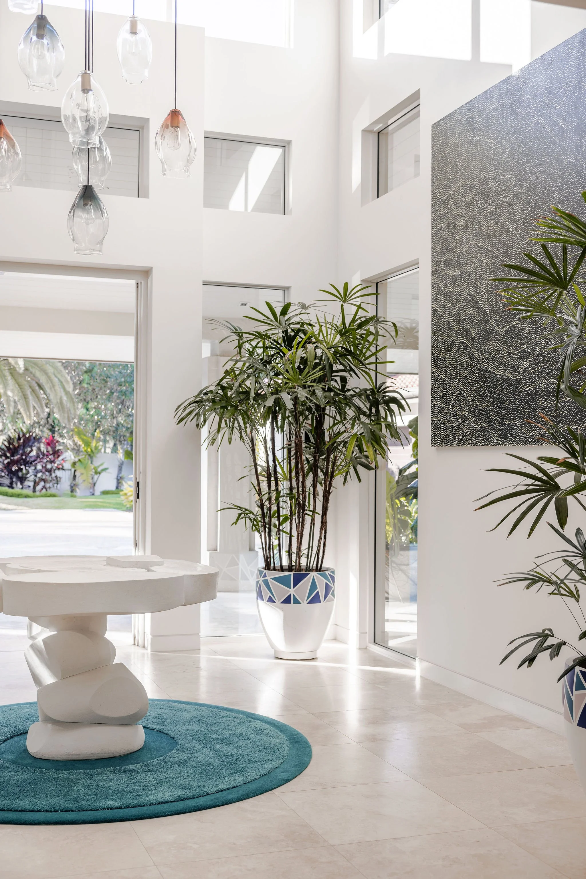 Bright modern interior with white walls and natural light, featuring a large potted green plant, a white artistic side table, a circular blue rug, and contemporary pendant lights.
