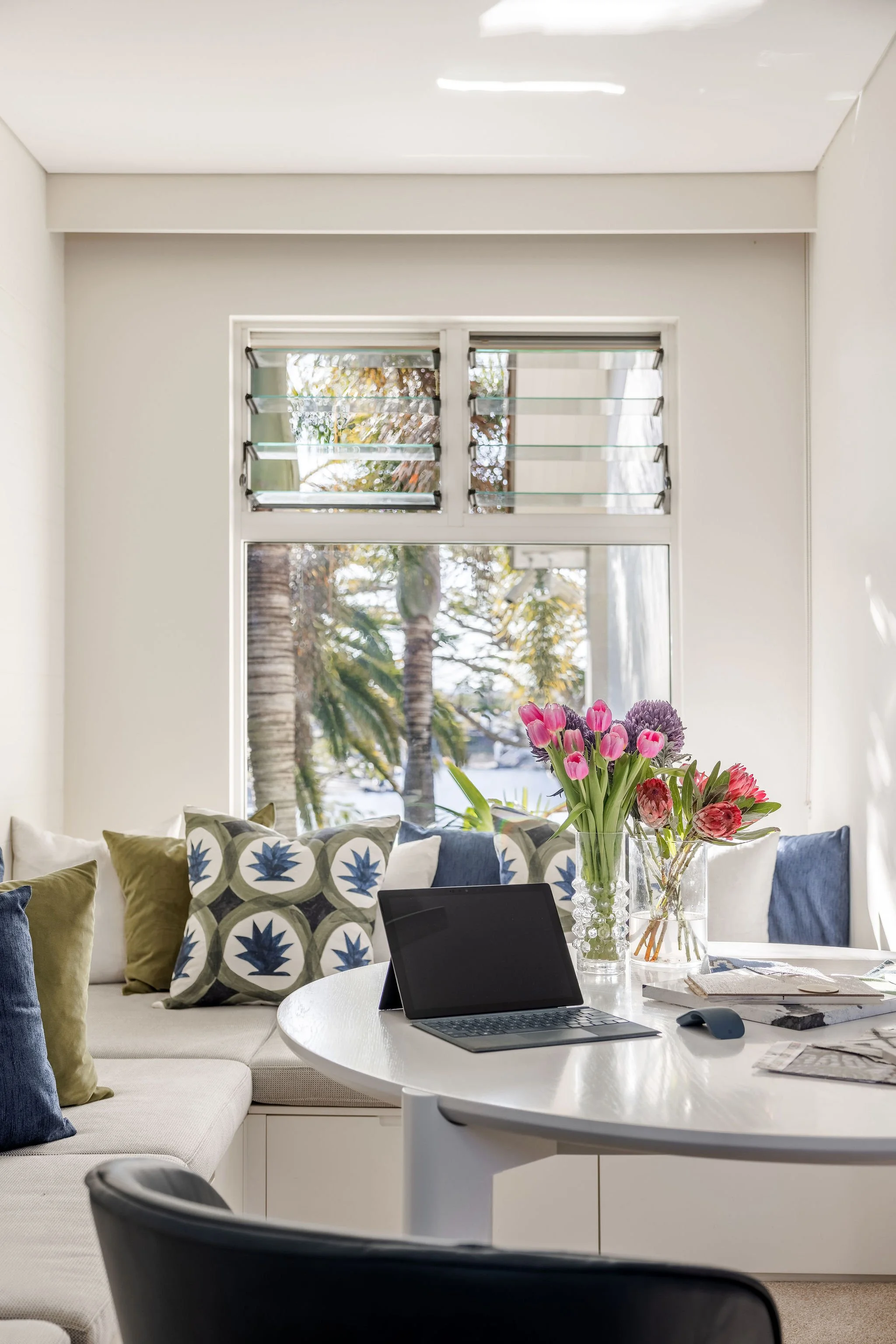 A Broadbeach Waters living room with a white wall, large window with roll-up shutters, floral arrangement on a white round table, and sectional sofa with patterned and plain pillows, laptop, mouse, and magazines on the table.