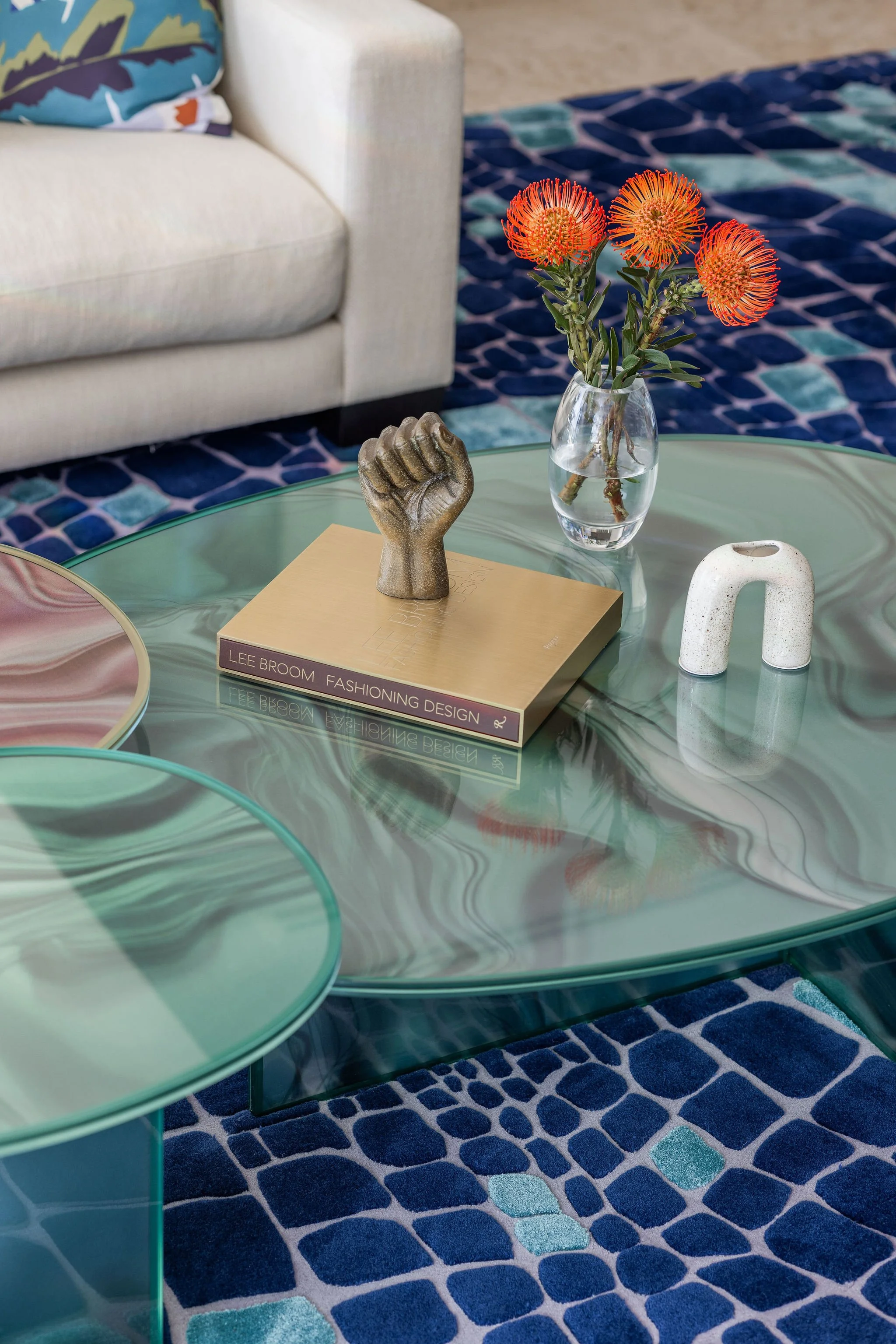 A living room with a glass coffee table, a vase with orange flowers, a beige sofa with a colorful pillow, a patterned rug, and decorative objects on the table.