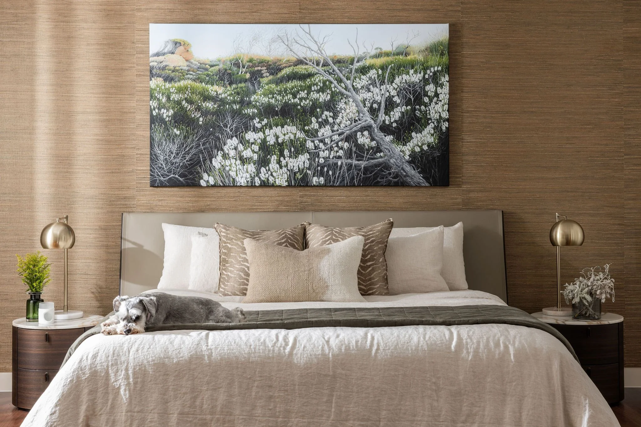A Gold Caost bedroom with a bed, decorative pillows, a dog lying on the bed, two bedside tables with lamps, a plant, a book, and a large framed painting of nature hanging on the wall