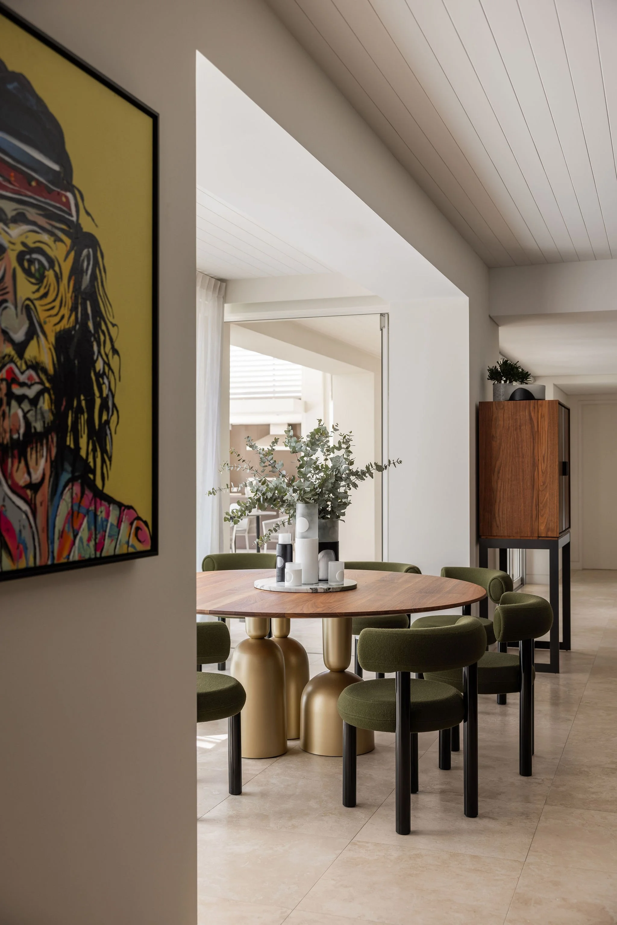 Modern dining room with green chairs around a round wooden table, decorated with a vase of eucalyptus and candles, with a large window in the background and artwork on the wall.