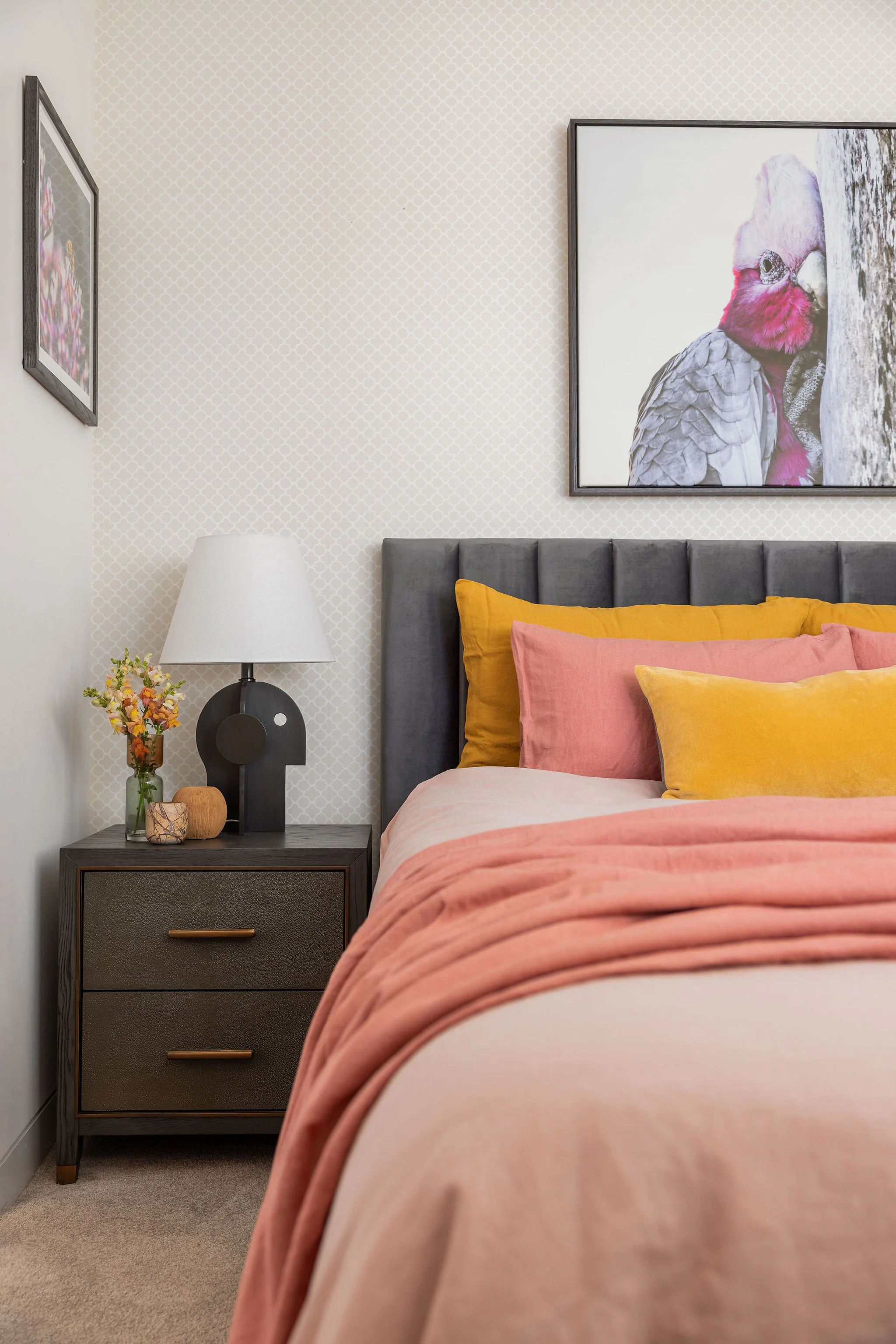 A bedroom with a bed, pillows in yellow, pink, and beige, a dark gray headboard, and a nightstand with a lamp, small flowers in a glass vase, and decorative items. There is a large framed photo of a pink and gray bird on the patterned wall.