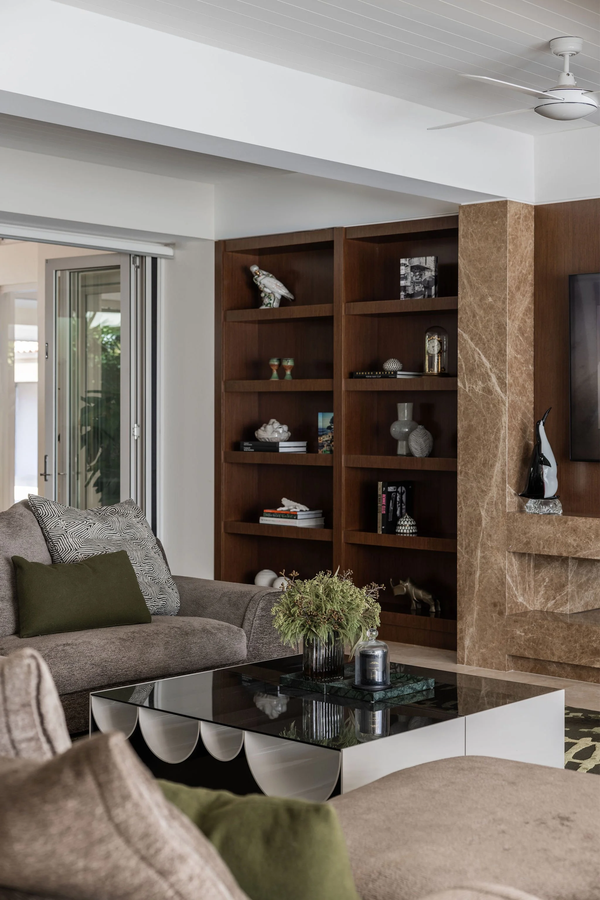 Living room with a gray sofa, green and black pillows, a decorative plant on a black and white glass coffee table, a wooden bookshelf with various ornaments, and a marble like wall section. Part of a sliding glass door is visible on the left, and a c