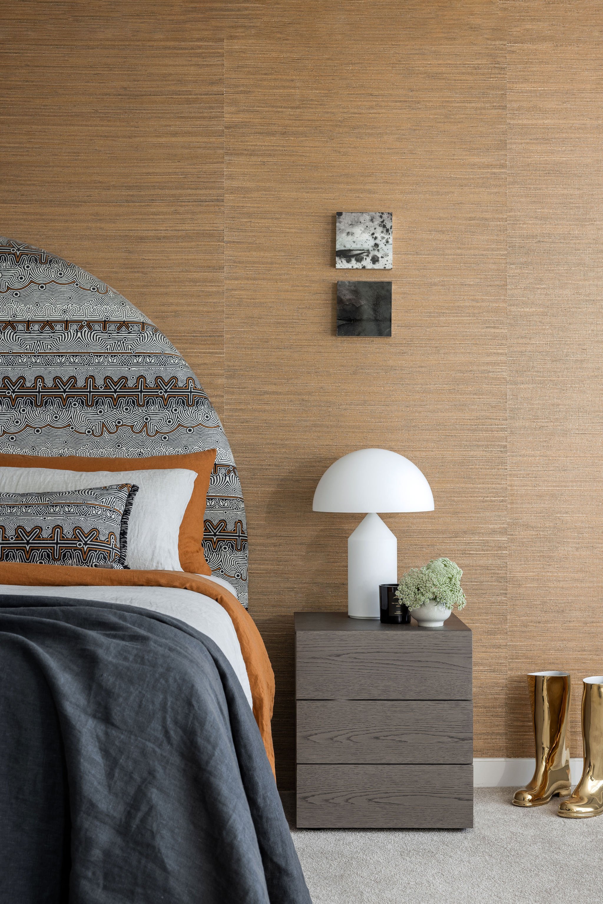 Bedroom with a wooden nightstand, a white table lamp, and decorative items, including a small clock and a plant, next to a bed with patterned bedding and a gray blanket. Gold-colored boot-shaped decor is on the carpeted floor, and the wall behind is 