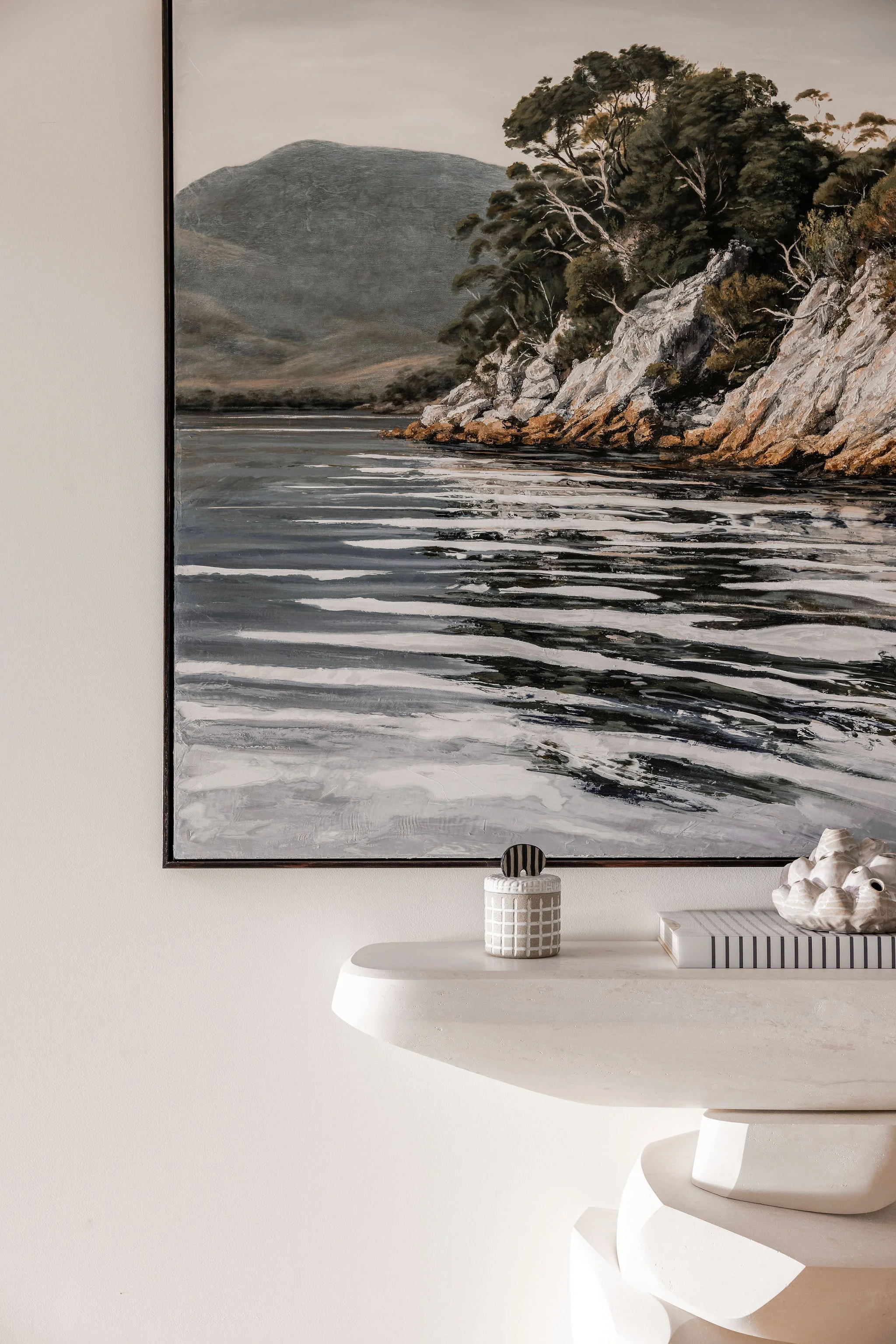 A minimalist interior wall features a large landscape painting of a river with a rocky cliff and trees, hung above a white, curved shelf decorated with a white geometric vase and a small sculpture, with part of a white chair visible in the foreground