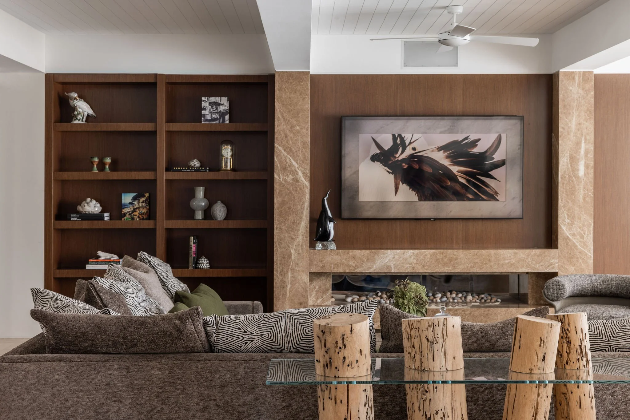 A luxurious Broadbeach Waters living room with a wooden built-in bookshelf filled with decorative objects, a large framed picture of feathers on the wall, a ceiling fan, and a cozy seating area with patterned cushions and wooden stools.