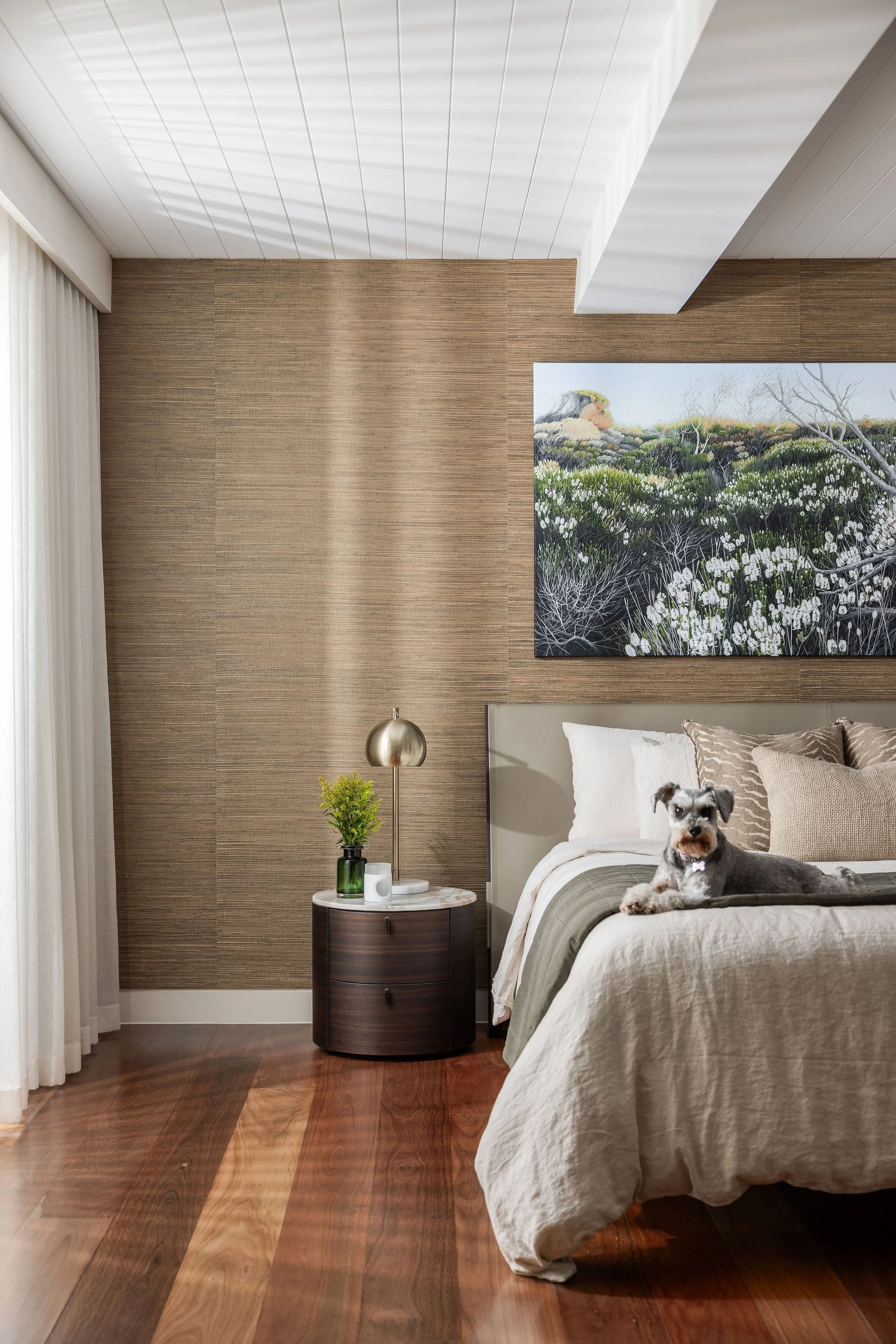 A cozy bedroom with wooden flooring, a bed with beige bedding, a nightstand with a small green plant, a lamp, and white curtains. There is a large painting of a landscape hanging on the wall and a small dog sitting on the bed.