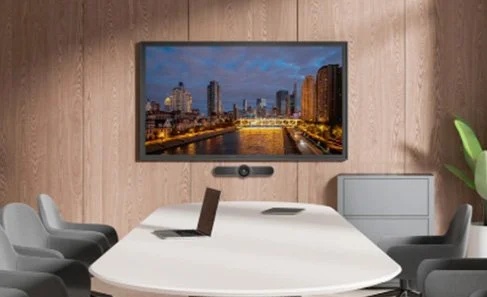 Conference room with a large monitor displaying a cityscape at dusk, a laptop open on a white oval table, and six gray chairs.