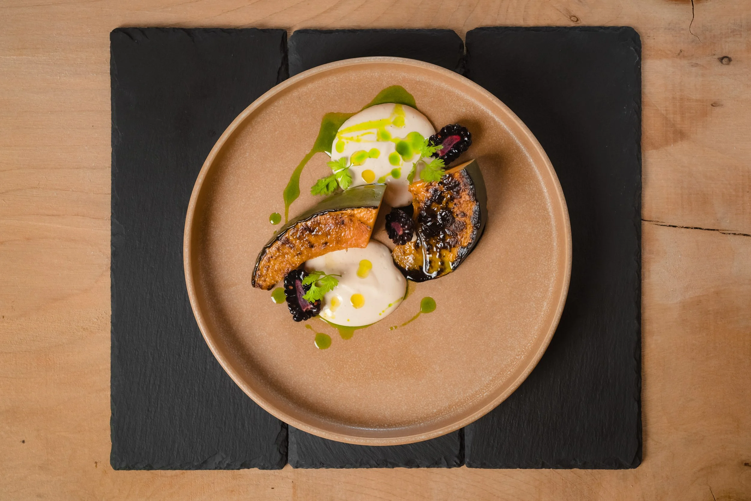 A gourmet dish featuring roasted eggplant slices, garnished with herbs and blackberries, served with dollops of white sauce and green oil drizzle on a beige plate, placed on a black stone slab on a wooden table.