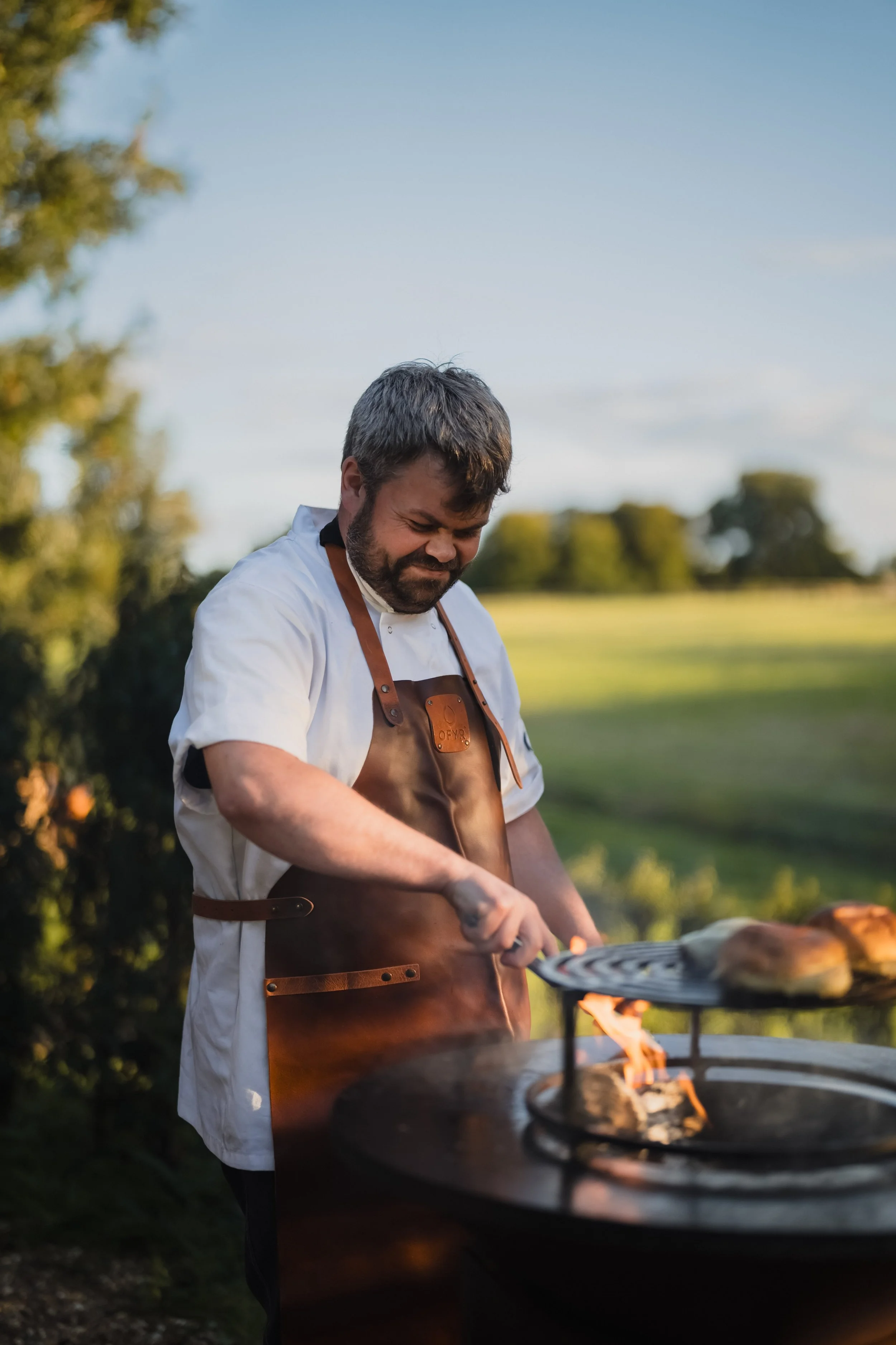 A man wearing a white shirt and brown apron cooking food on a grill outdoors in a scenic rural setting during sunset.