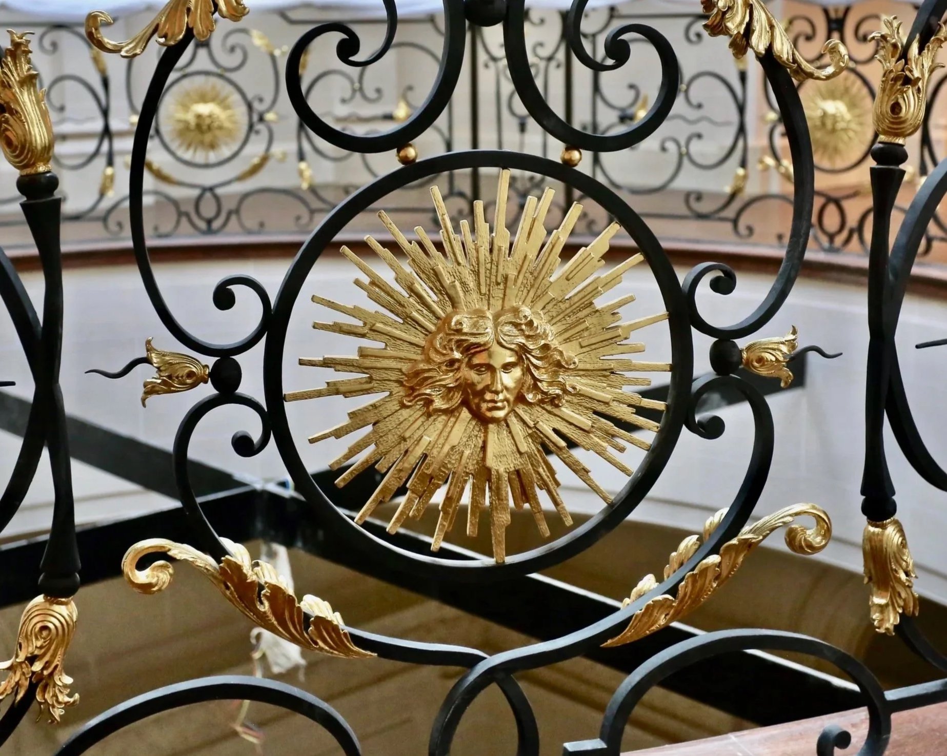 Close-up of a decorative wrought iron gate or fence with intricate black scrollwork, gold leaf accents, and a central golden sun with a patient's face surrounded by sun rays.