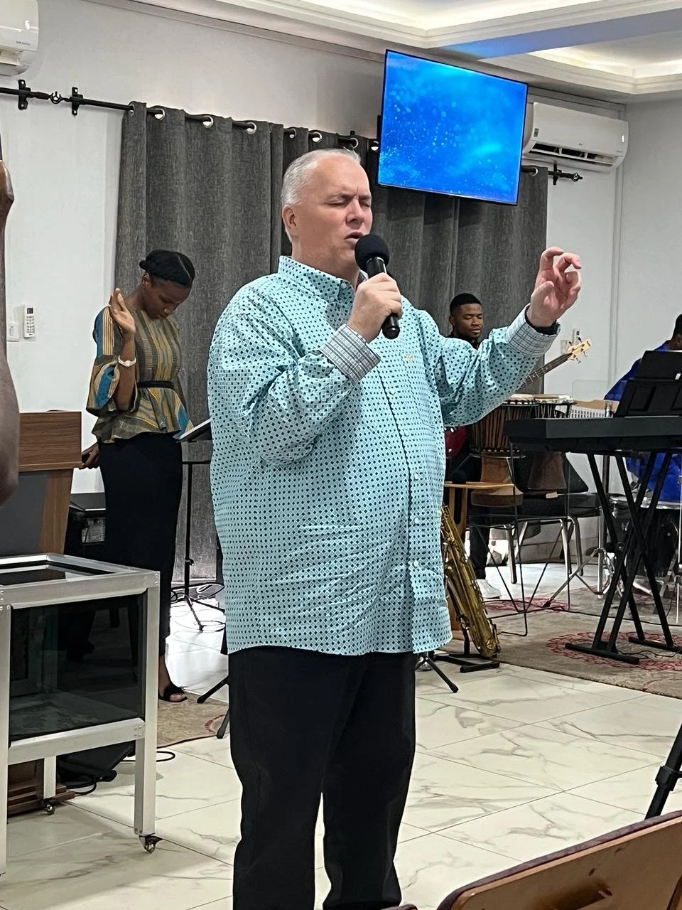 A man singing into a microphone with eyes closed, in a room with musicians in the background. A woman is standing with her head bowed, possibly praying or reflective. The setting appears to be a church or a music performance space with gray curtains,