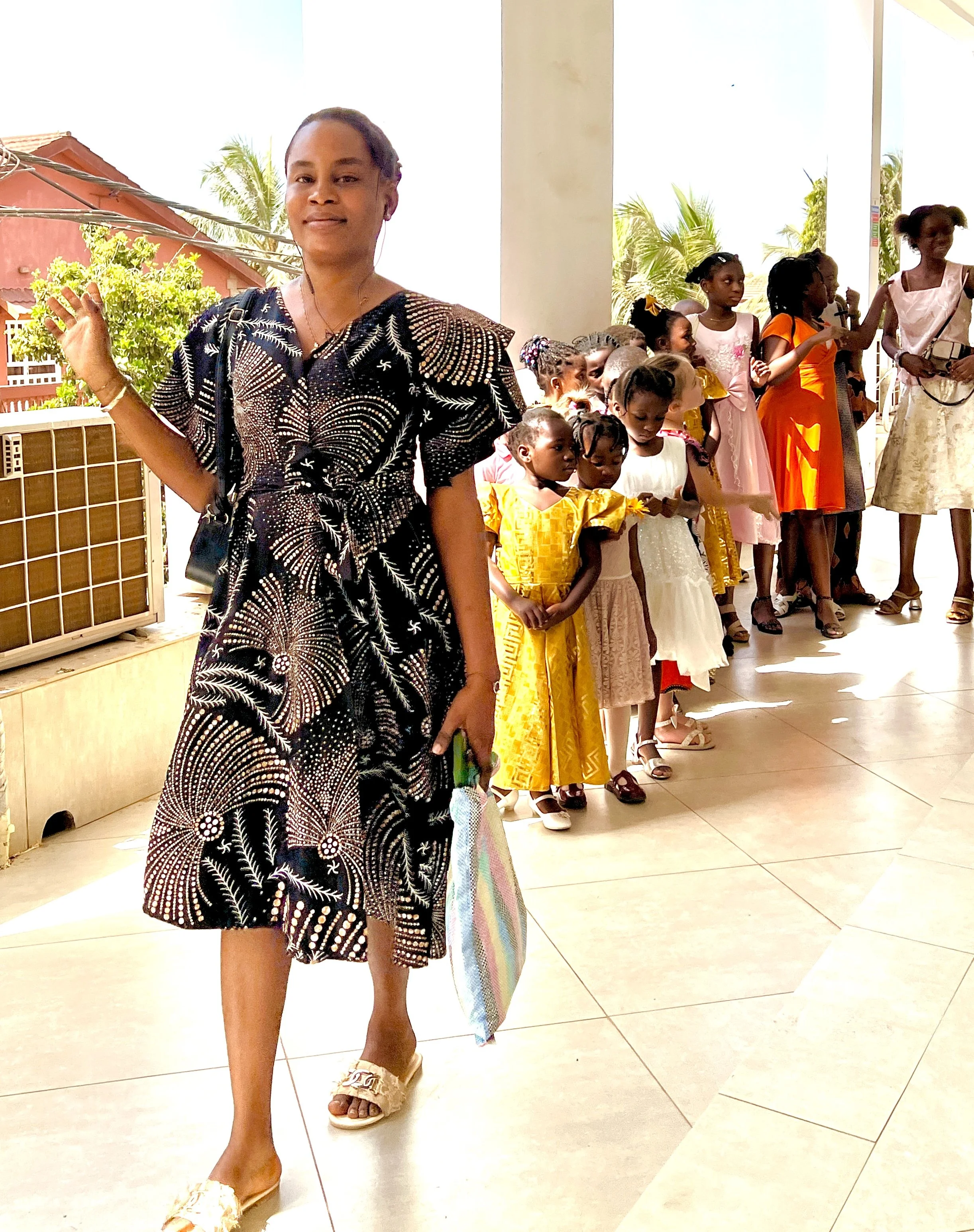 A woman in a black patterned dress stands on a tiled floor, waving and smiling. Behind her, a line of children and women in colorful dresses and casual shoes are standing, some with their hands raised or gesturing. Palm trees and red-roofed buildings