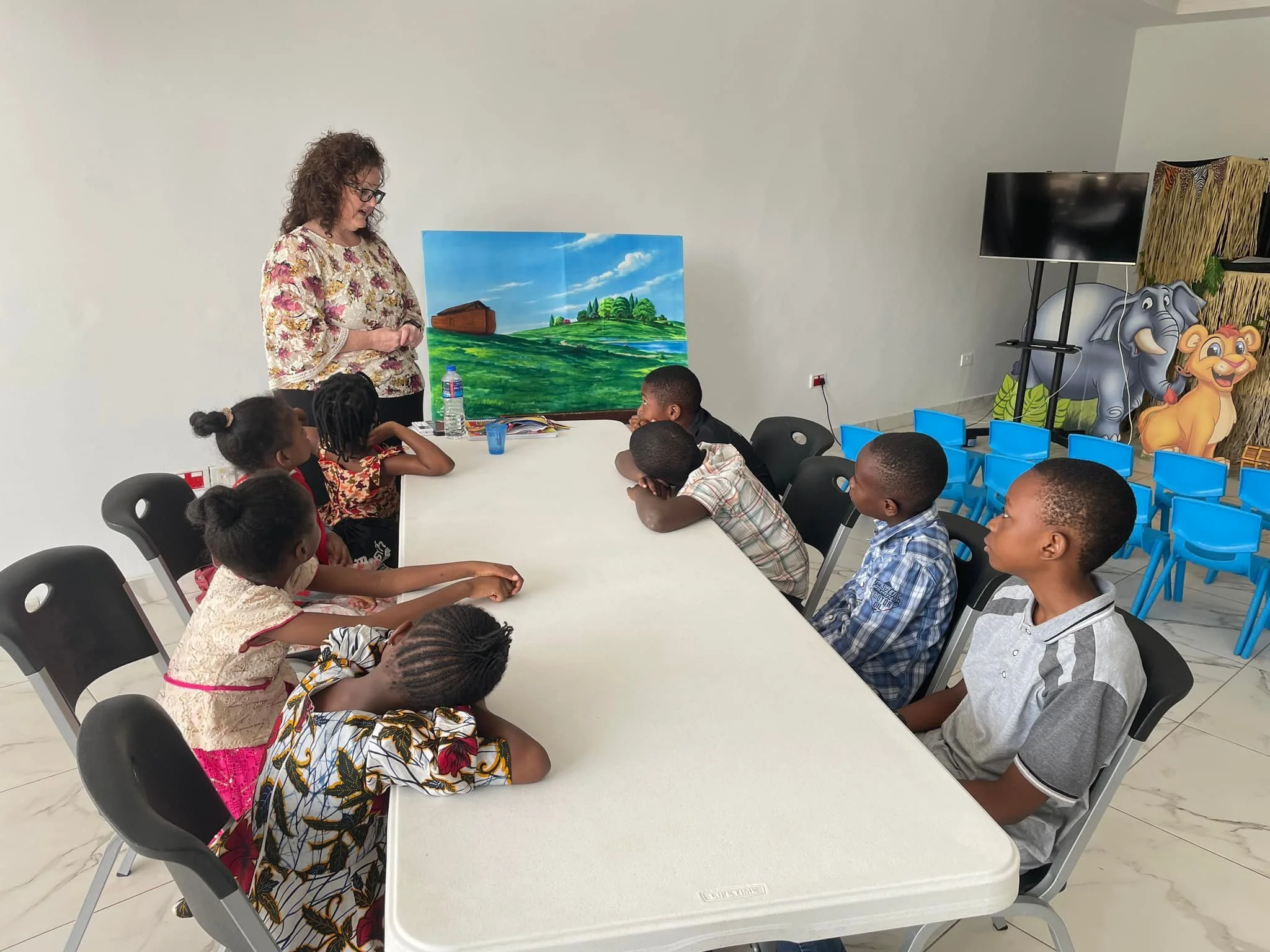 A woman teaching a group of children sitting at a white table in a classroom with colorful elephant and lion decorations, a TV, and a scenic landscape painting in the background.