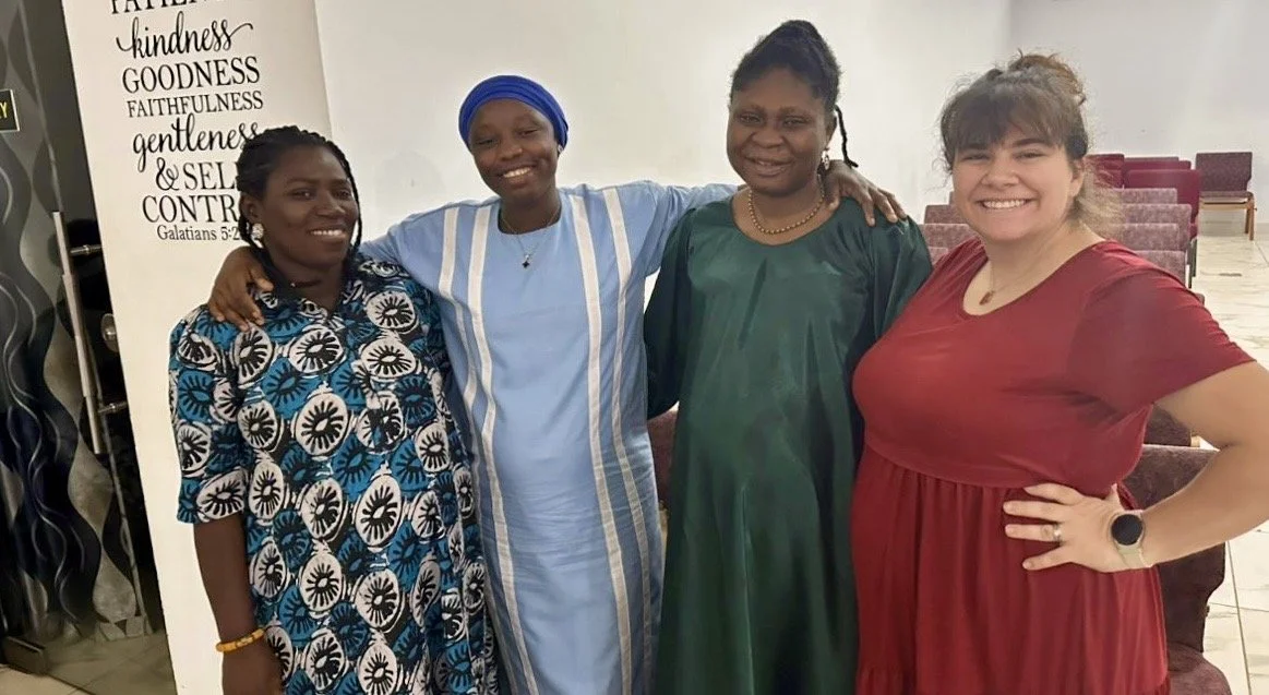 Four women standing close together indoors, smiling, with arm links around each other's shoulders.