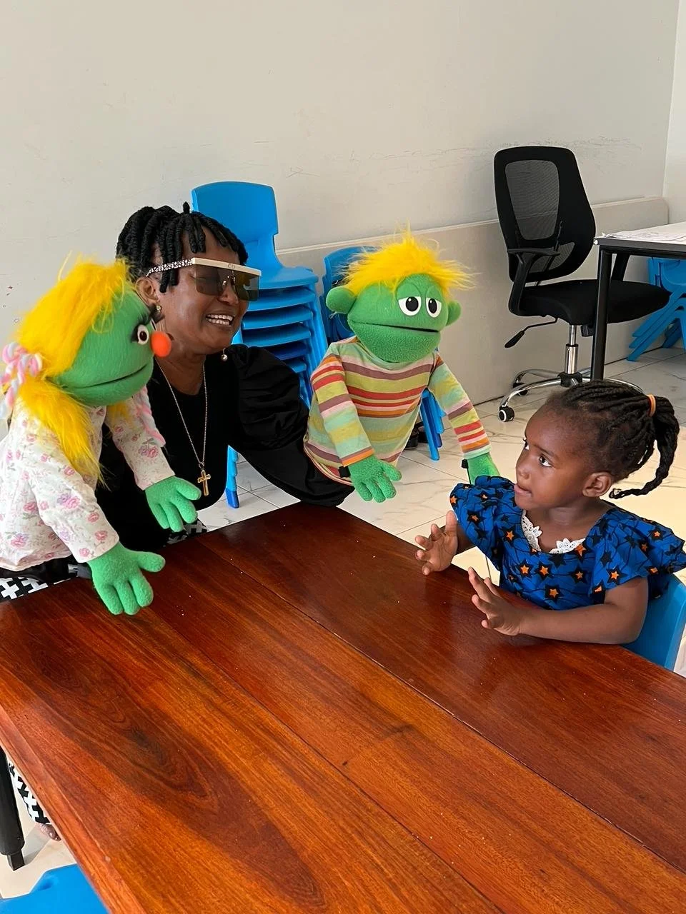 A young girl sitting at a table engaging with two puppets dressed as colorful monsters with green faces, bright yellow hair, and striped shirts. An adult woman, wearing sunglasses and a cross necklace, is also present, smiling and interacting.