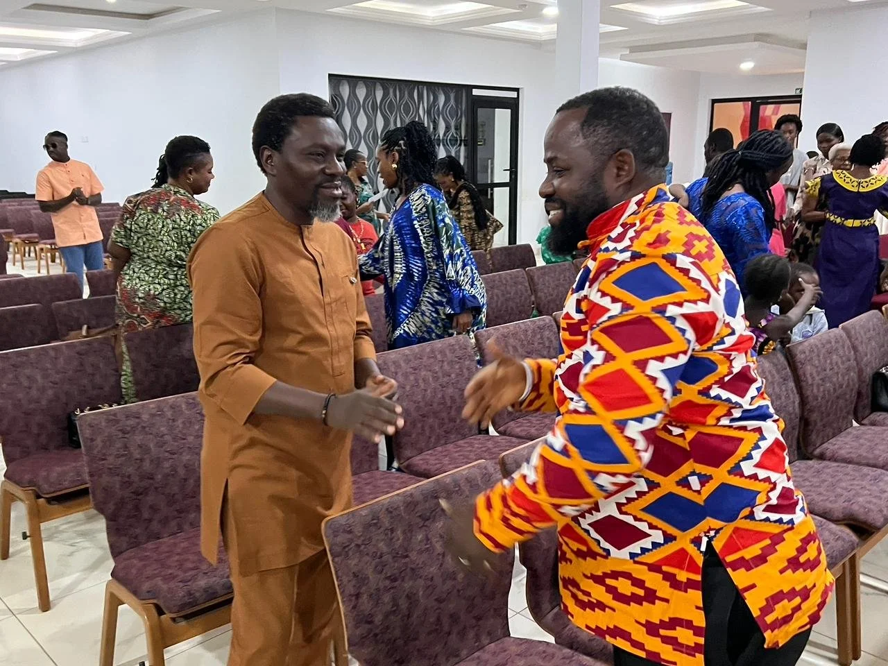 Two men are greeting each other with a handshake and smiling. They are surrounded by other people in a large room with chairs. The man on the left is wearing a brown traditional outfit, and the man on the right is wearing a colorful patterned shirt.