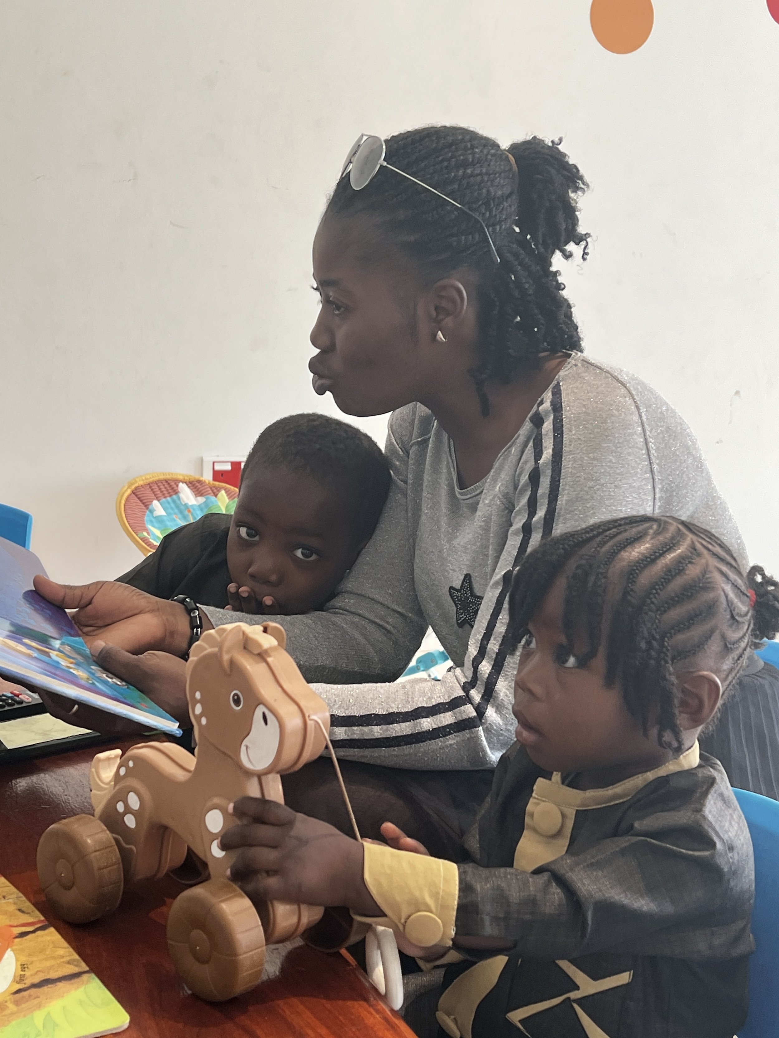 A woman and three children sitting around a table playing with toys, including a horse figurine. The woman is wearing sunglasses on her head and a gray shirt with black stripes. The children have braided hair, one with a yellow jacket and the other w