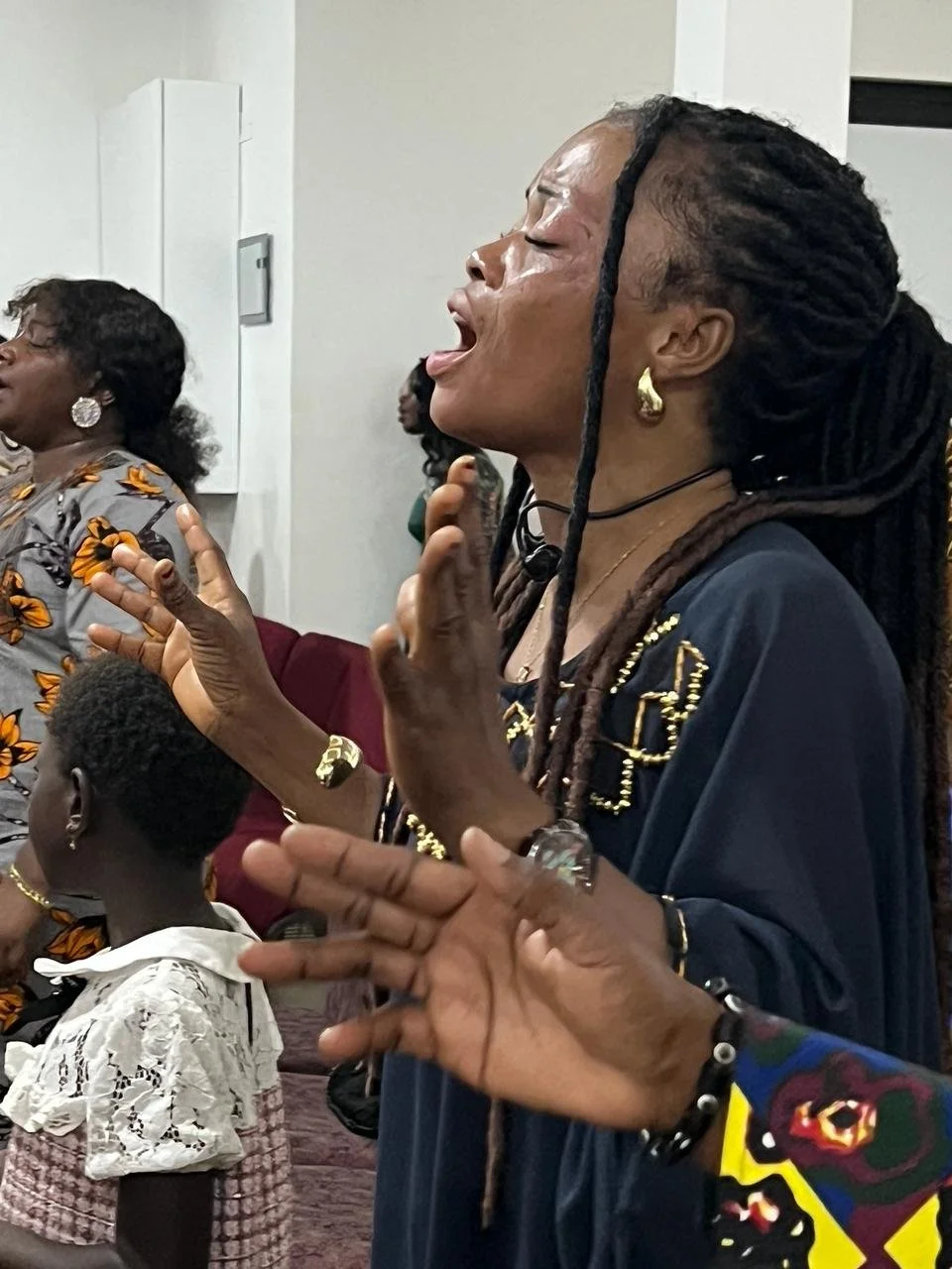 People gathered in prayer or worship, with eyes closed and hands raised, in a room with white walls.