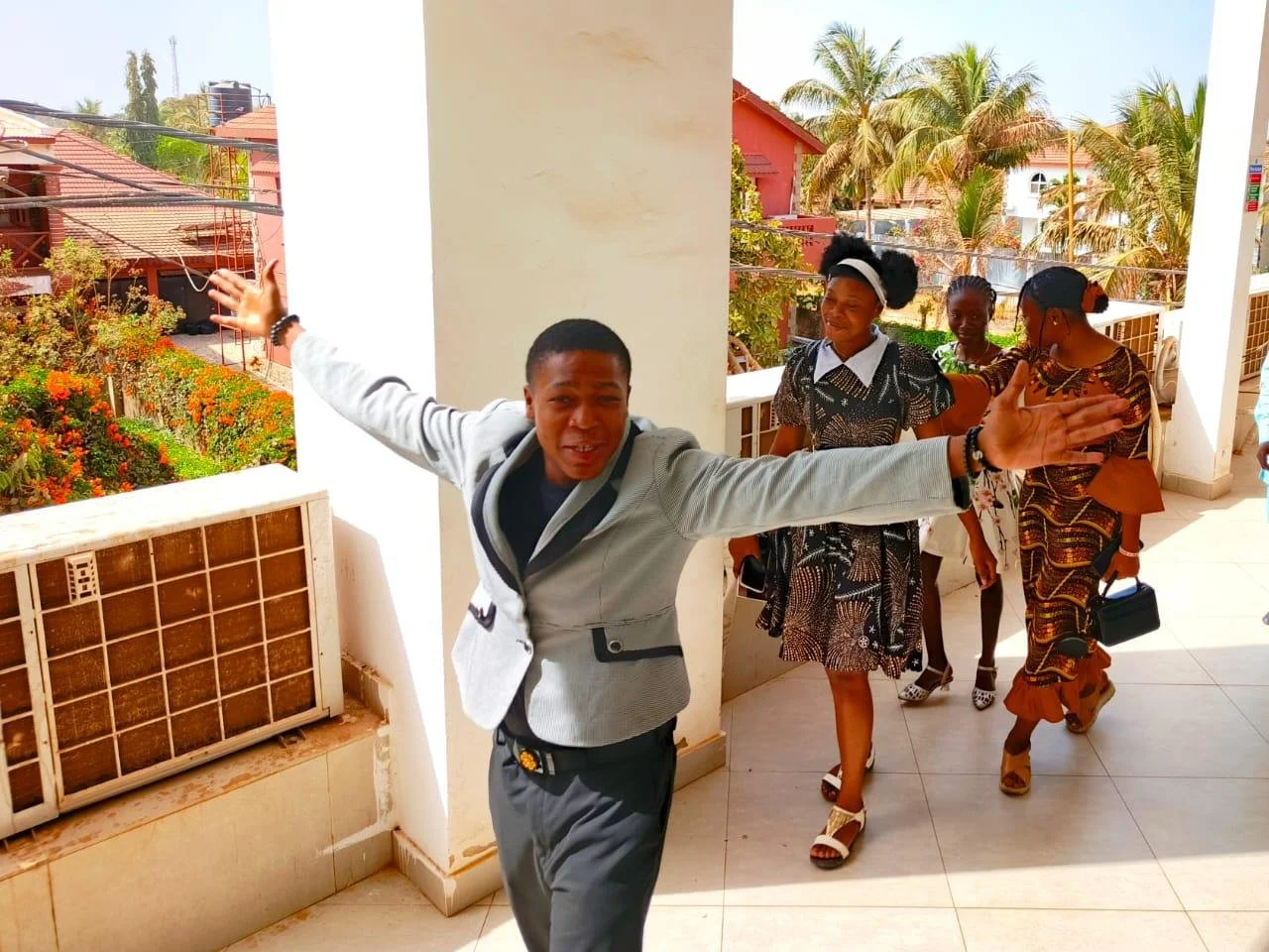 A young man with his arms outstretched, smiling, standing on a balcony, with several women walking behind him in a sunny outdoor setting with trees and houses.