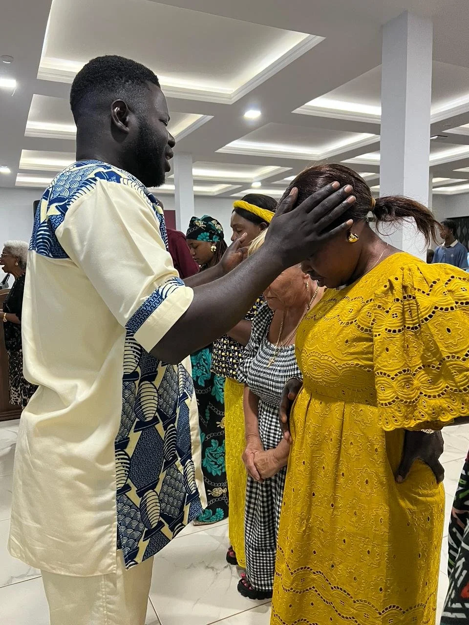 A man gently touching a woman's forehead during prayer at a religious gathering. The woman is wearing a yellow dress, and several women are standing with their heads bowed in the background.