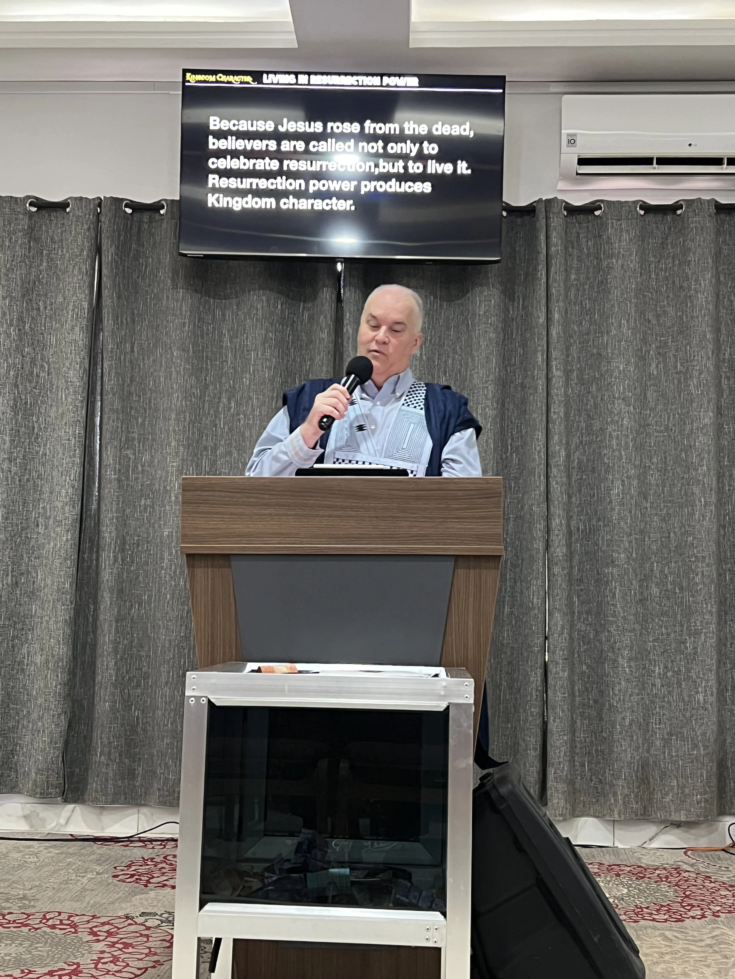 A speaker at a podium giving a speech, with a large screen behind him displaying a message about resurrection power and spiritual beliefs. The setting appears to be a conference or church event.