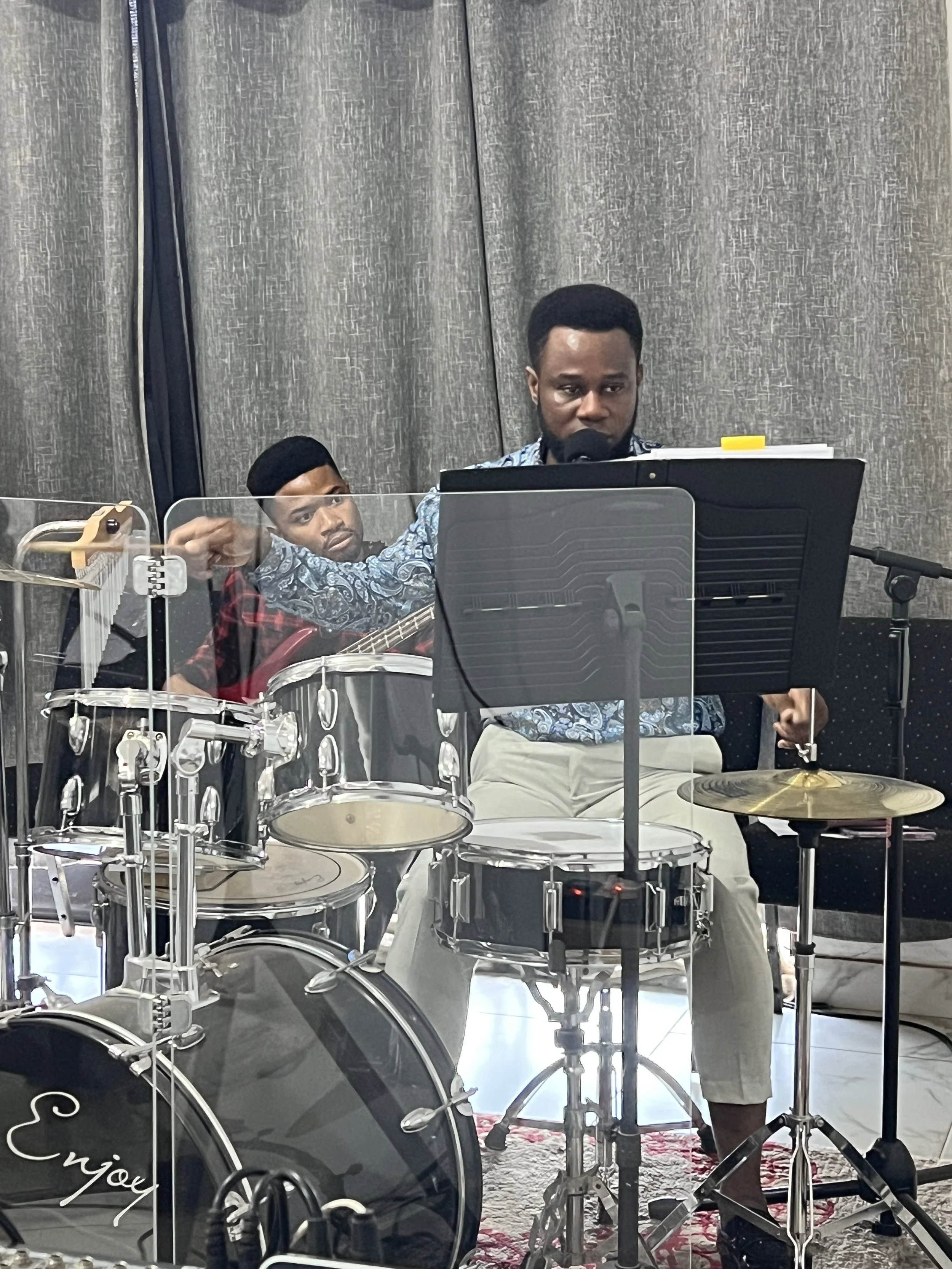 Two men sitting behind music stands and a drum set. One man has a guitar. The setting appears to be a practice room or stage with gray curtains and a patterned rug.