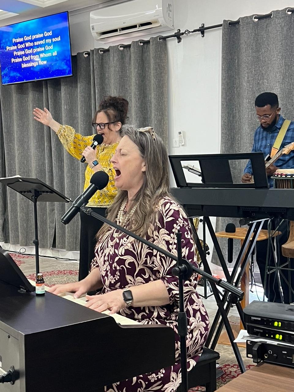 A woman playing the piano and singing into a microphone, a woman in a yellow top singing into a microphone, and a man playing the guitar in a room with gray curtains and a wall-mounted TV displaying lyrics.