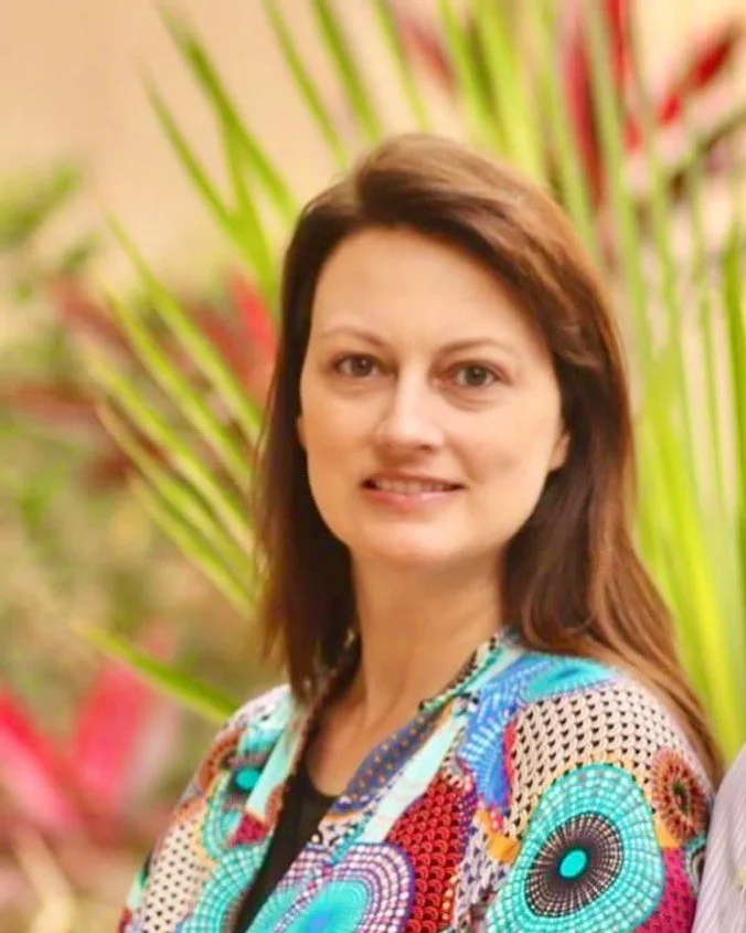 Woman with brown hair smiling outdoors, wearing colorful patterned clothing, with green and red plants in the background.