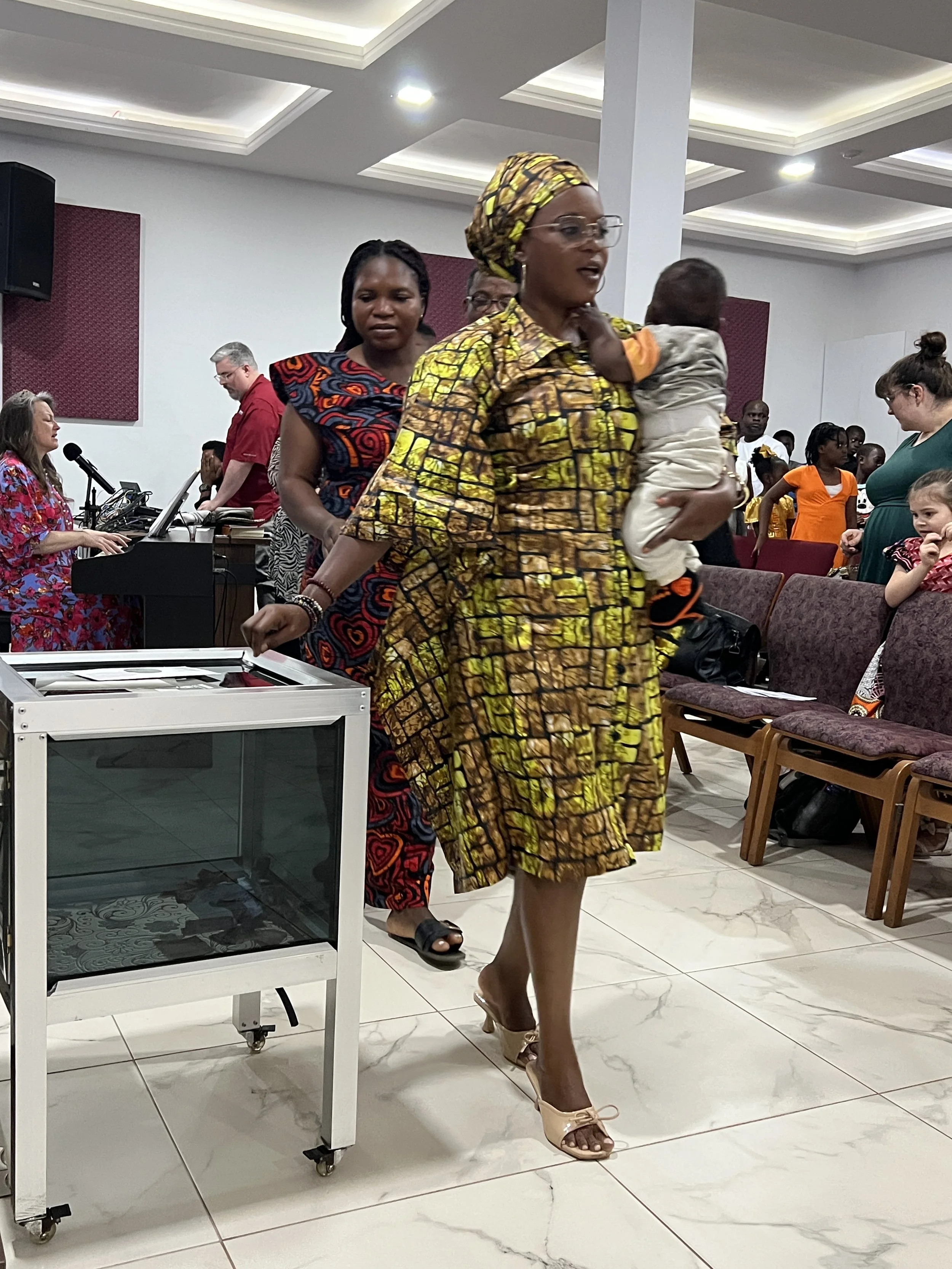 A woman in traditional African attire is walking in a crowded indoor space, holding a child on her shoulder. Behind her, a woman in a patterned dress and a man in red are visible, with several other people in the background. There is a piano laptop a