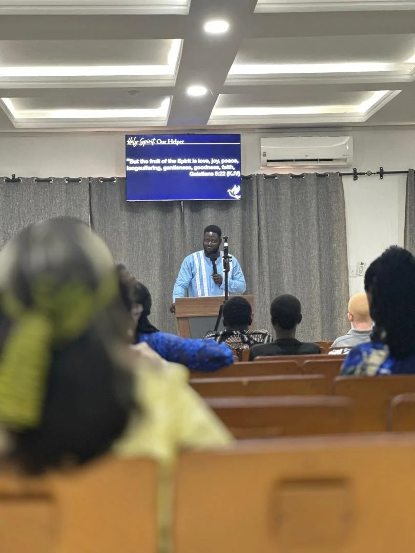 A man is speaking at a podium in front of an audience in a room with ceiling lights and gray curtains. A screen behind him displays a religious quote from Galatians 5:22-23. The audience members are seated and facing the speaker, including a woman with a yellow headscarf in the foreground.