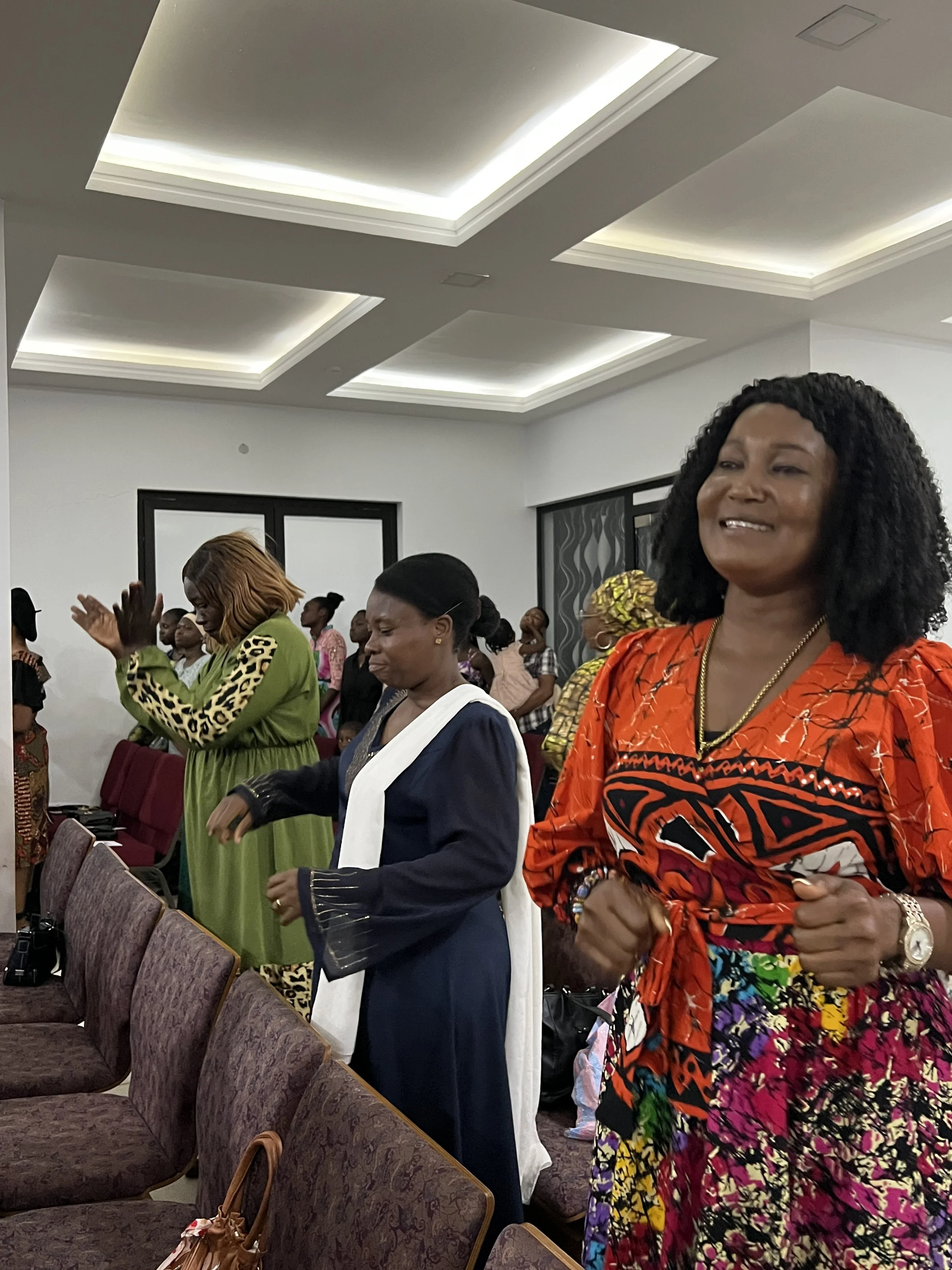 A group of women standing in prayer during a religious service or gathering inside a church or conference hall, with some with eyes closed, hands raised or clasped.