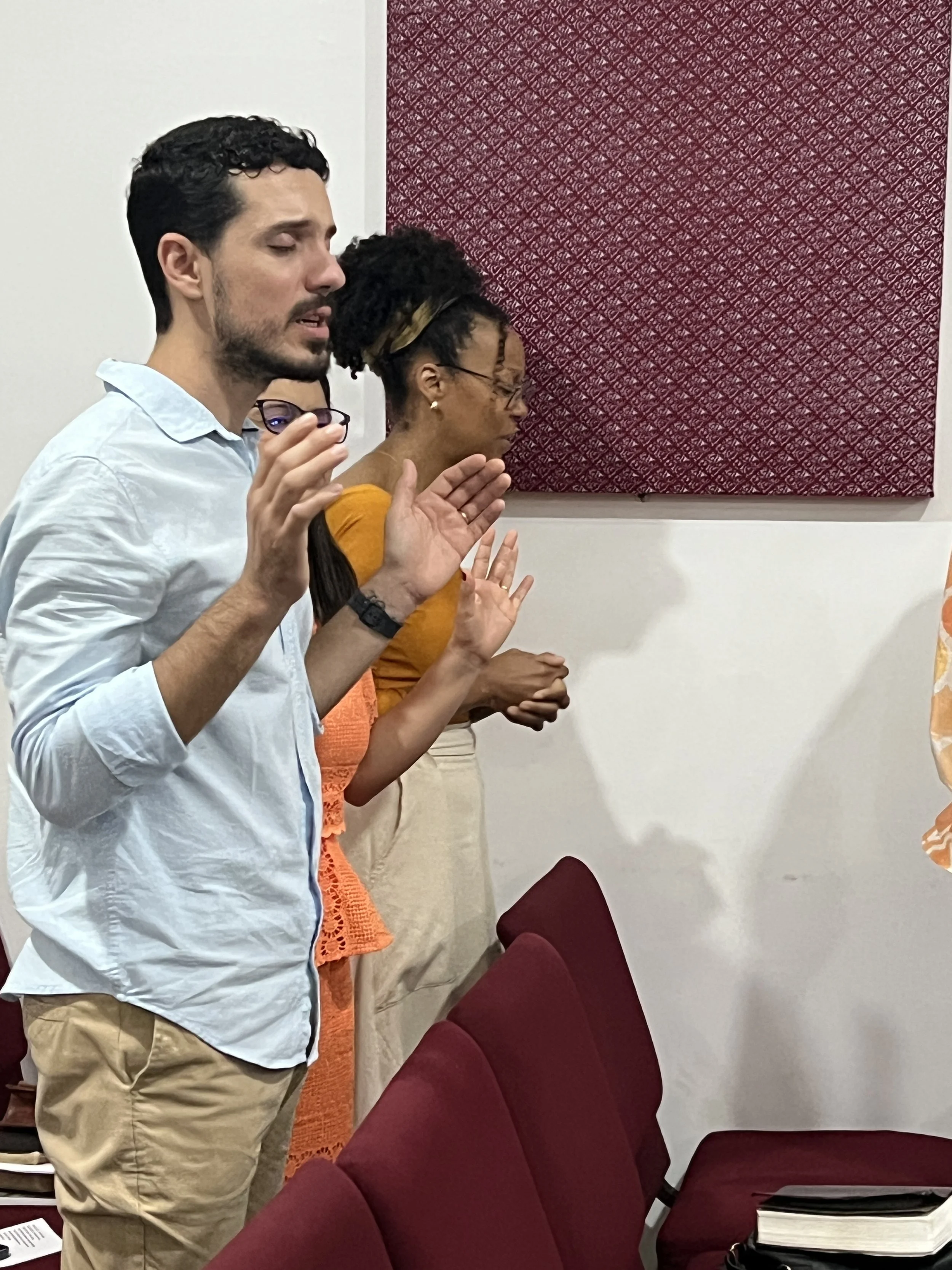 People standing with eyes closed and hands raised, possibly praying in a room with red chairs and a patterned artwork on the wall.