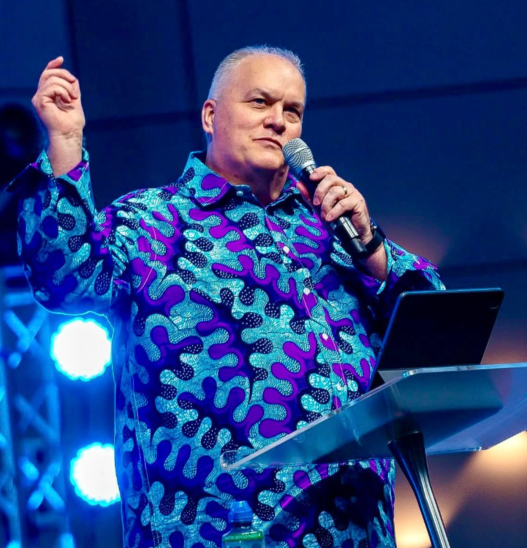 A man with short gray hair speaking into a microphone on stage, wearing a colorful patterned shirt, with stage lights in the background.