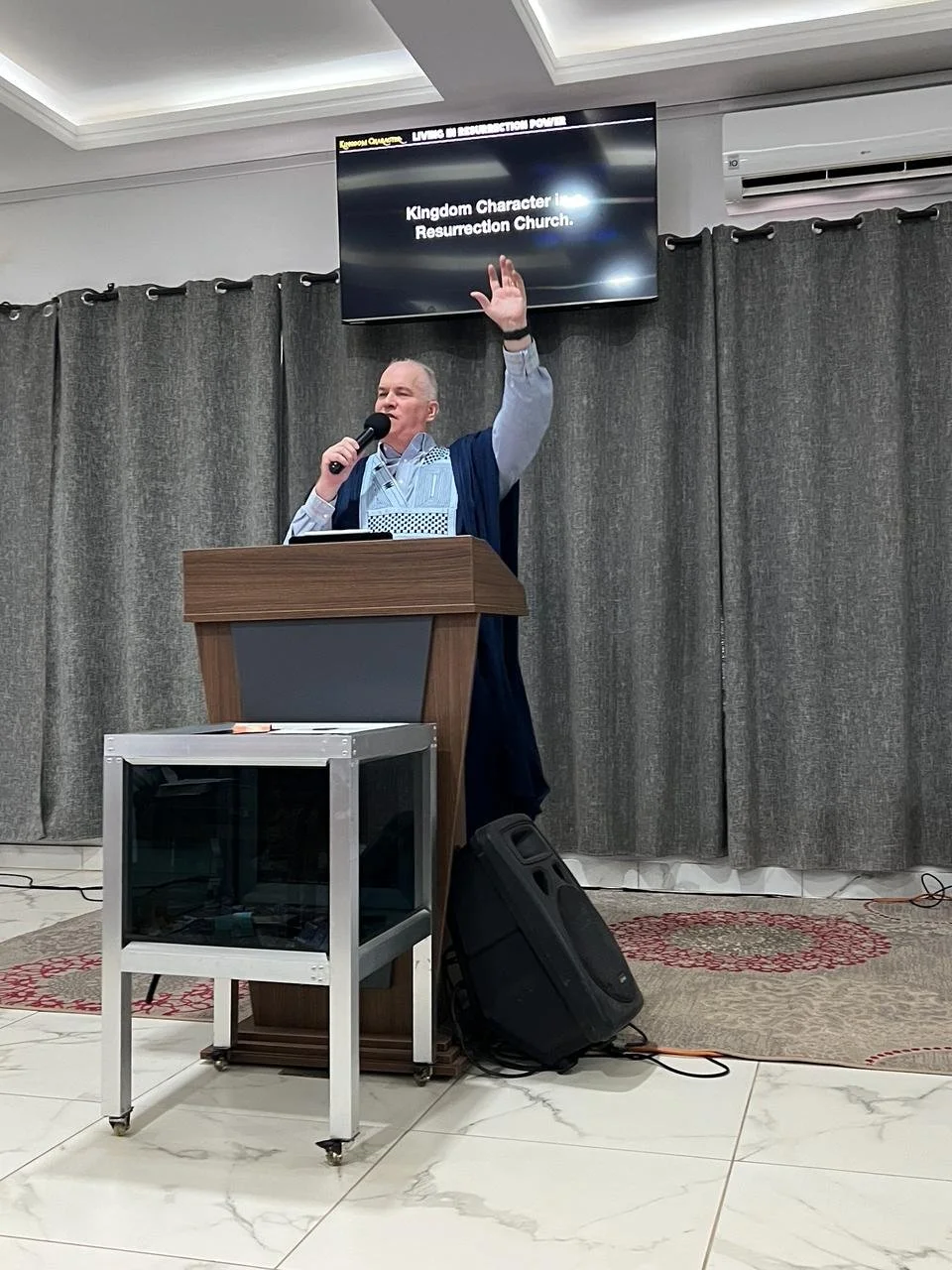 A man is speaking at a podium with a microphone in front of gray curtains. There is a television screen above him displaying text related to a church service. A speaker is on the floor next to the podium, and a small white and glass table is in front