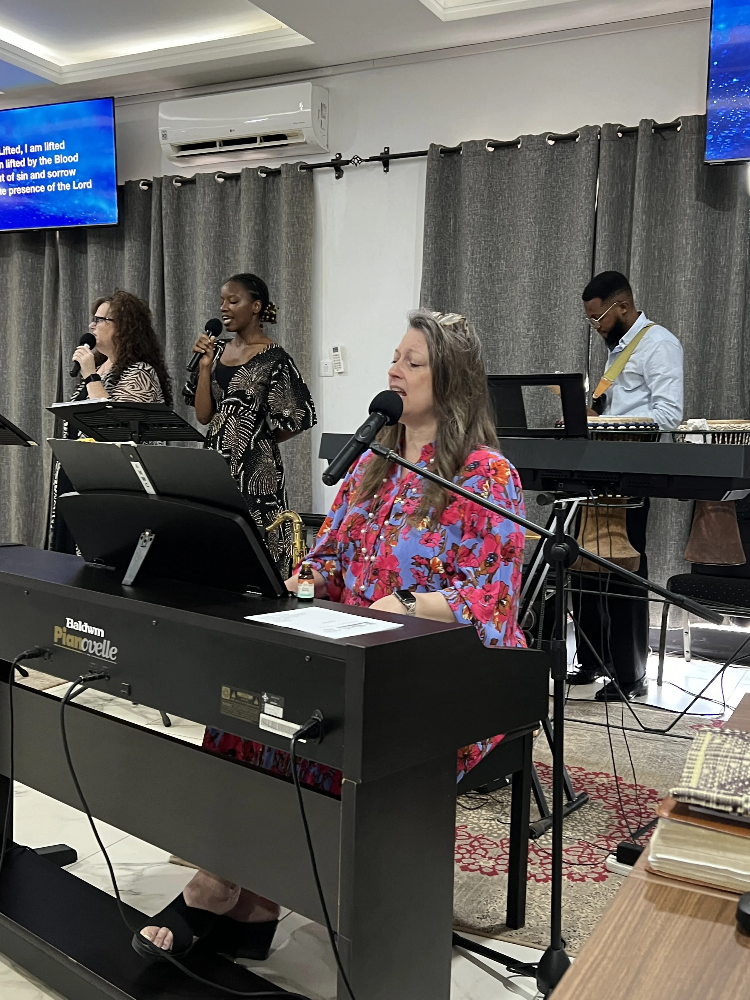 A group of people singing and playing musical instruments on stage during a church or community service, with a woman in a floral shirt playing a keyboard at the front.