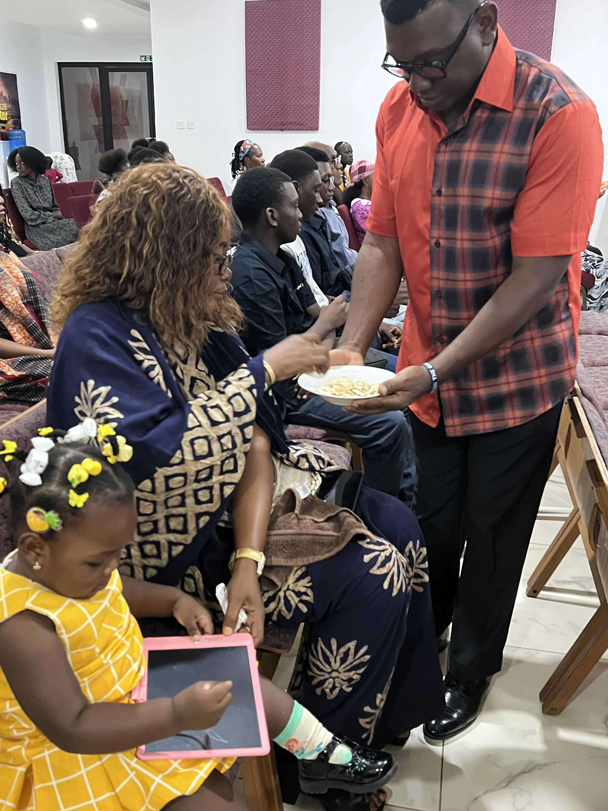 A man in a red and black shirt is handing out snacks to a woman in a blue and gold traditional dress, who is sitting with a group of people in an indoor setting. A young girl in a yellow dress is sitting nearby, playing with a tablet.