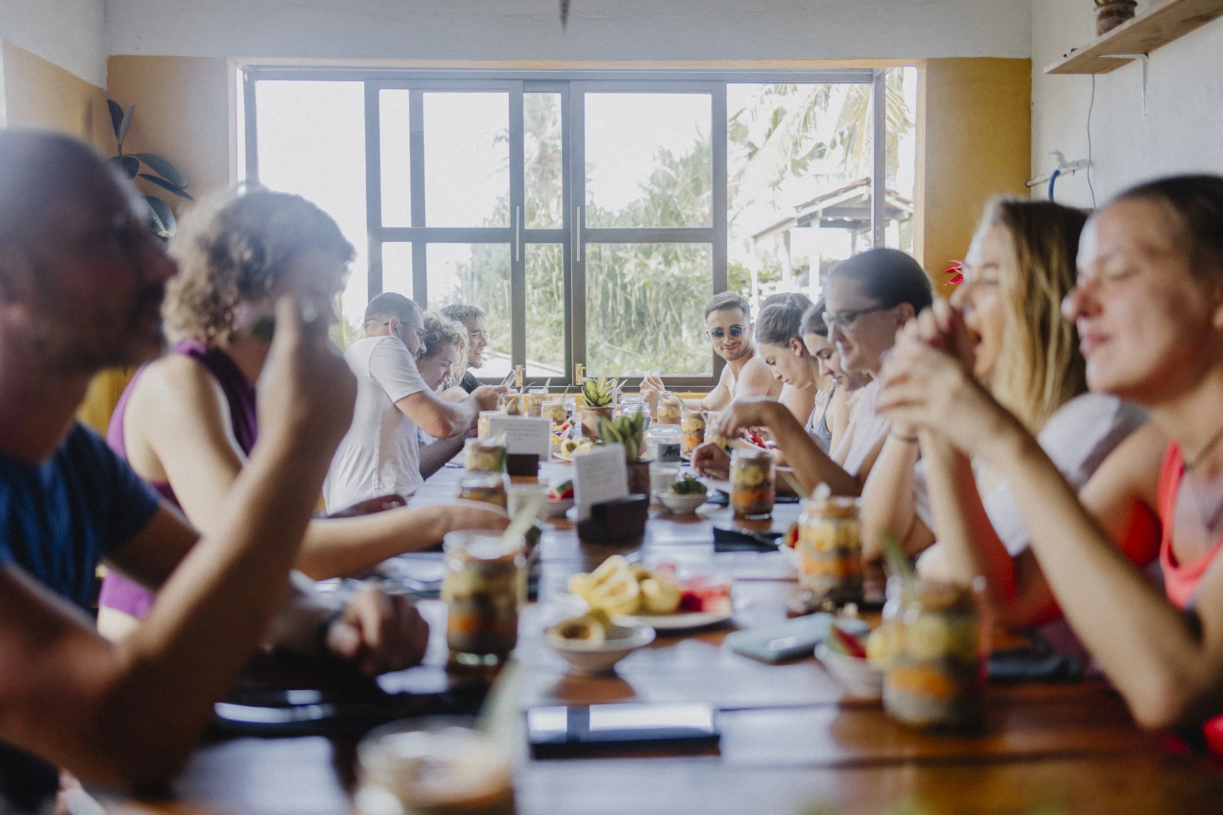 People sitting at a long dining table enjoying a meal in a bright room with large windows and plants outside.